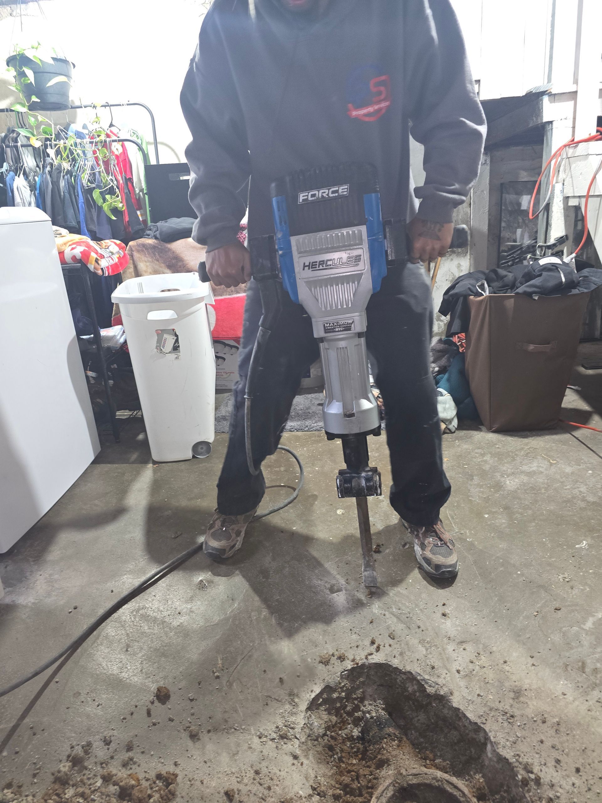 A person in a gray hooded sweatshirt operates a jackhammer to break up a concrete floor indoors.