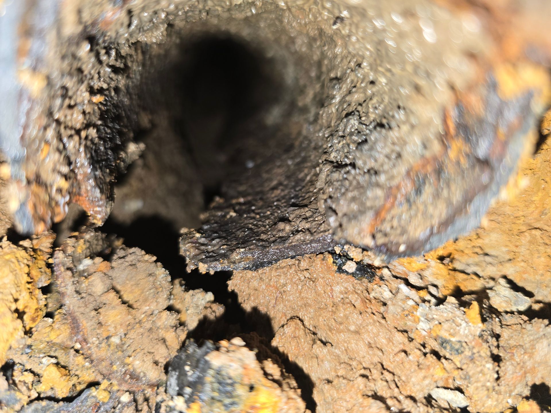 A close-up view looking into a corroded, debris-filled metal pipe embedded in rocky, brown soil.