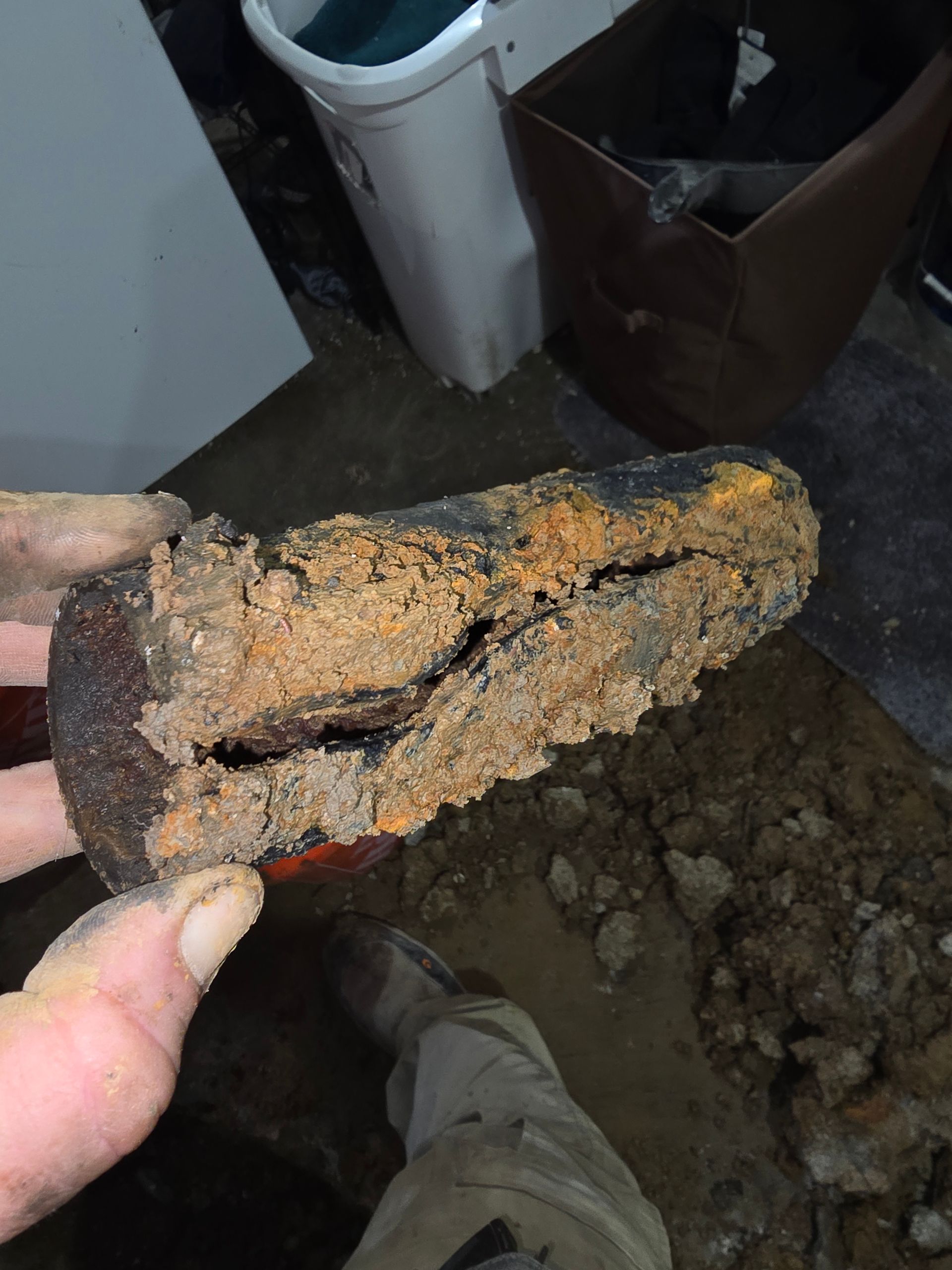 A close-up of a hand holding a piece of corroded pipe filled with orange sediment, with a construction site in the background.