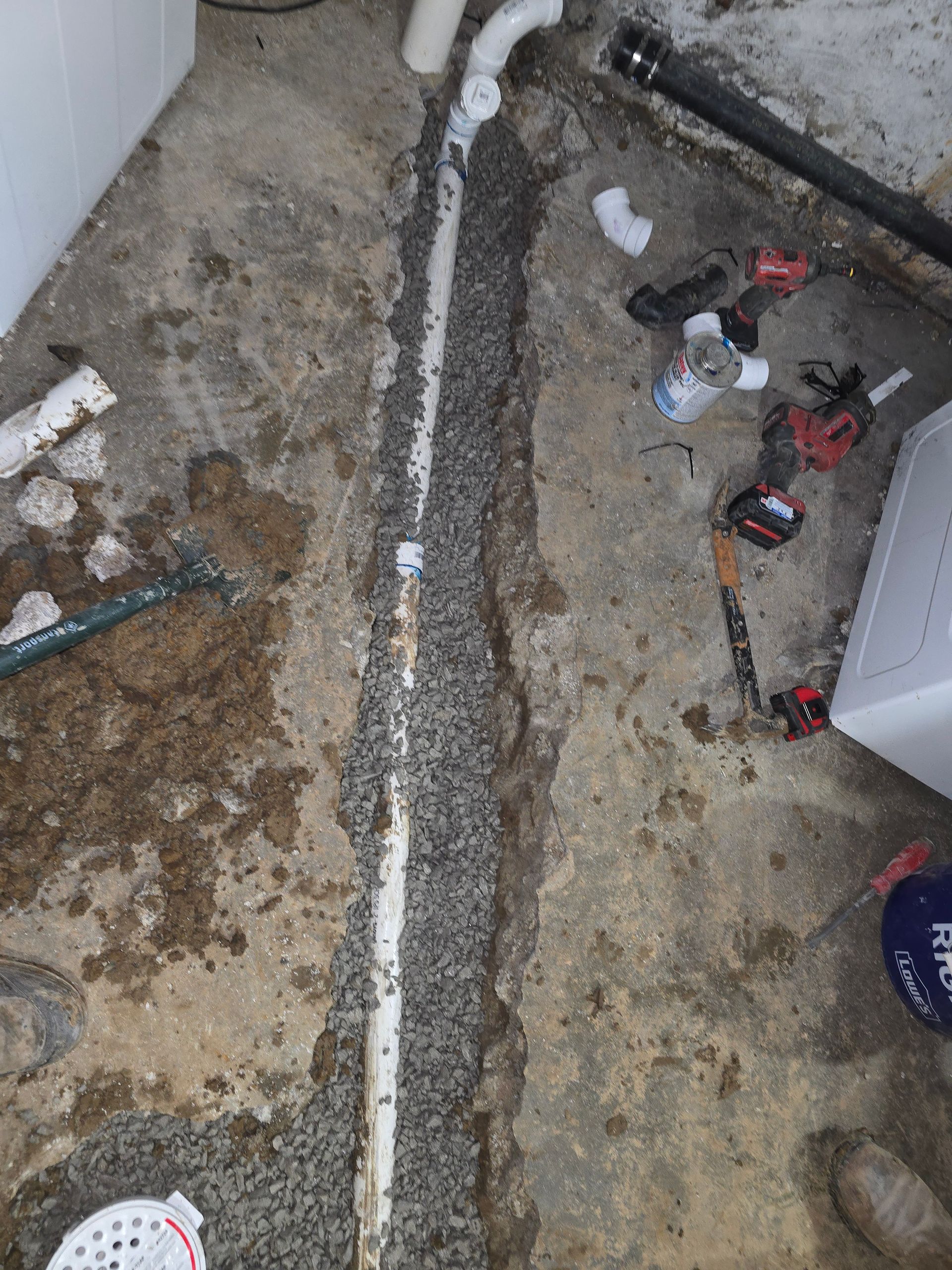 A white PVC drain pipe sits in a gravel-filled trench cut through a concrete basement floor, near plumbing tools.