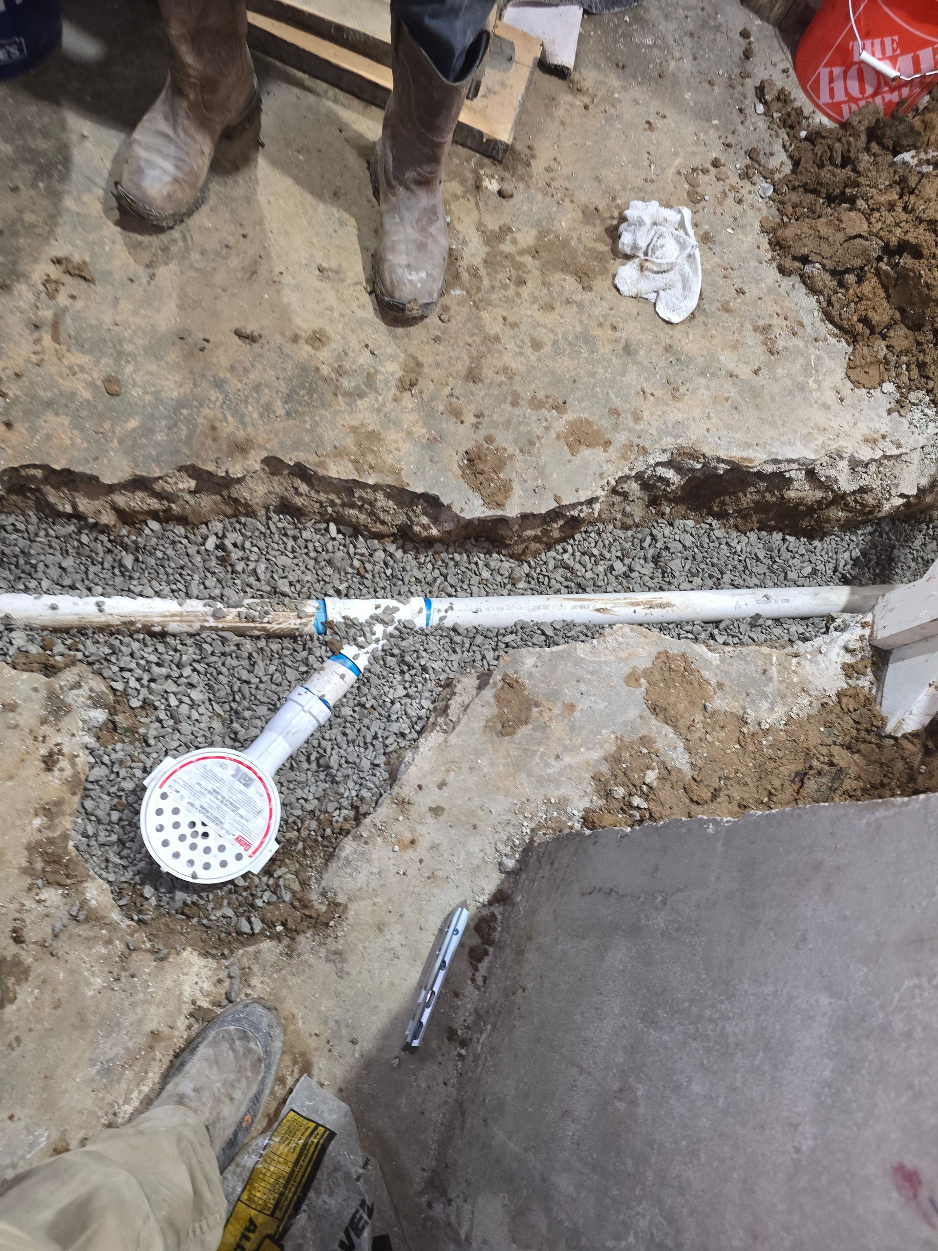 A white PVC drain pipe with a round floor grate is installed in a gravel-filled trench cut through a concrete floor.