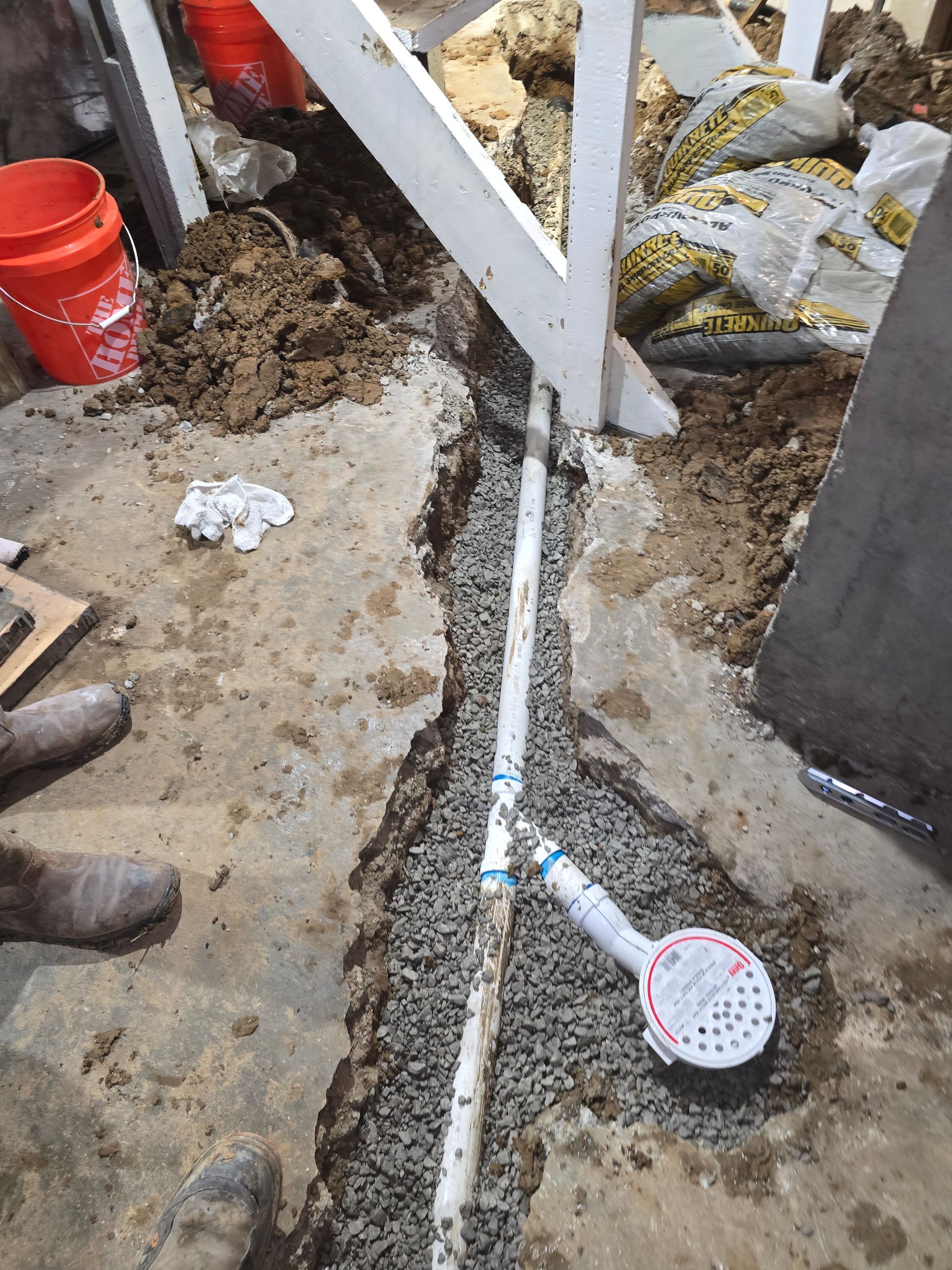 A white PVC pipe and floor drain installed in a trench filled with gravel through a broken concrete floor.