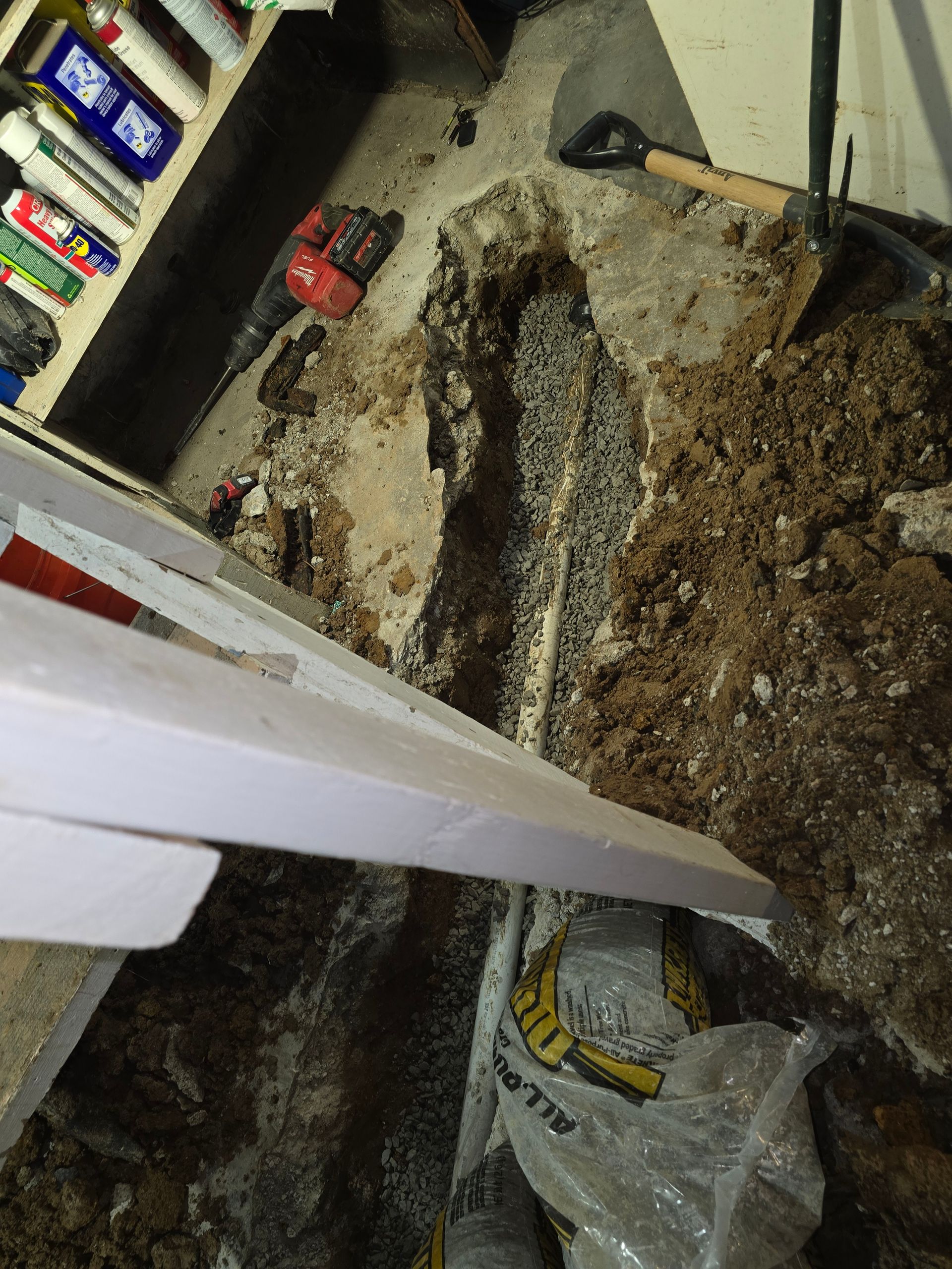 A trench dug through a concrete floor into the dirt, revealing a white pipe, with a shovel and tools nearby.
