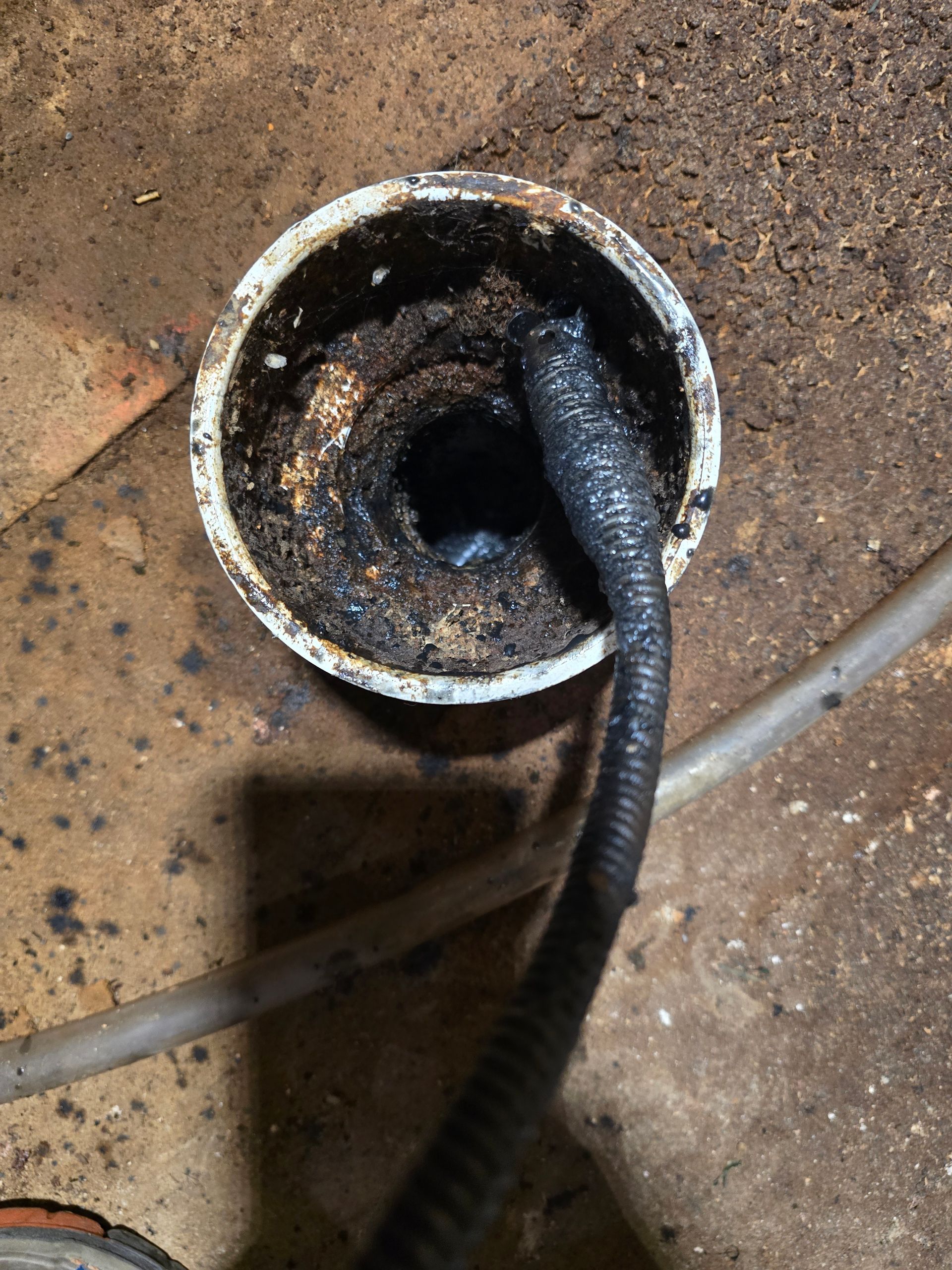 A plumbing snake cable inserted into a dark, debris-filled circular drain pipe on a dirt floor.