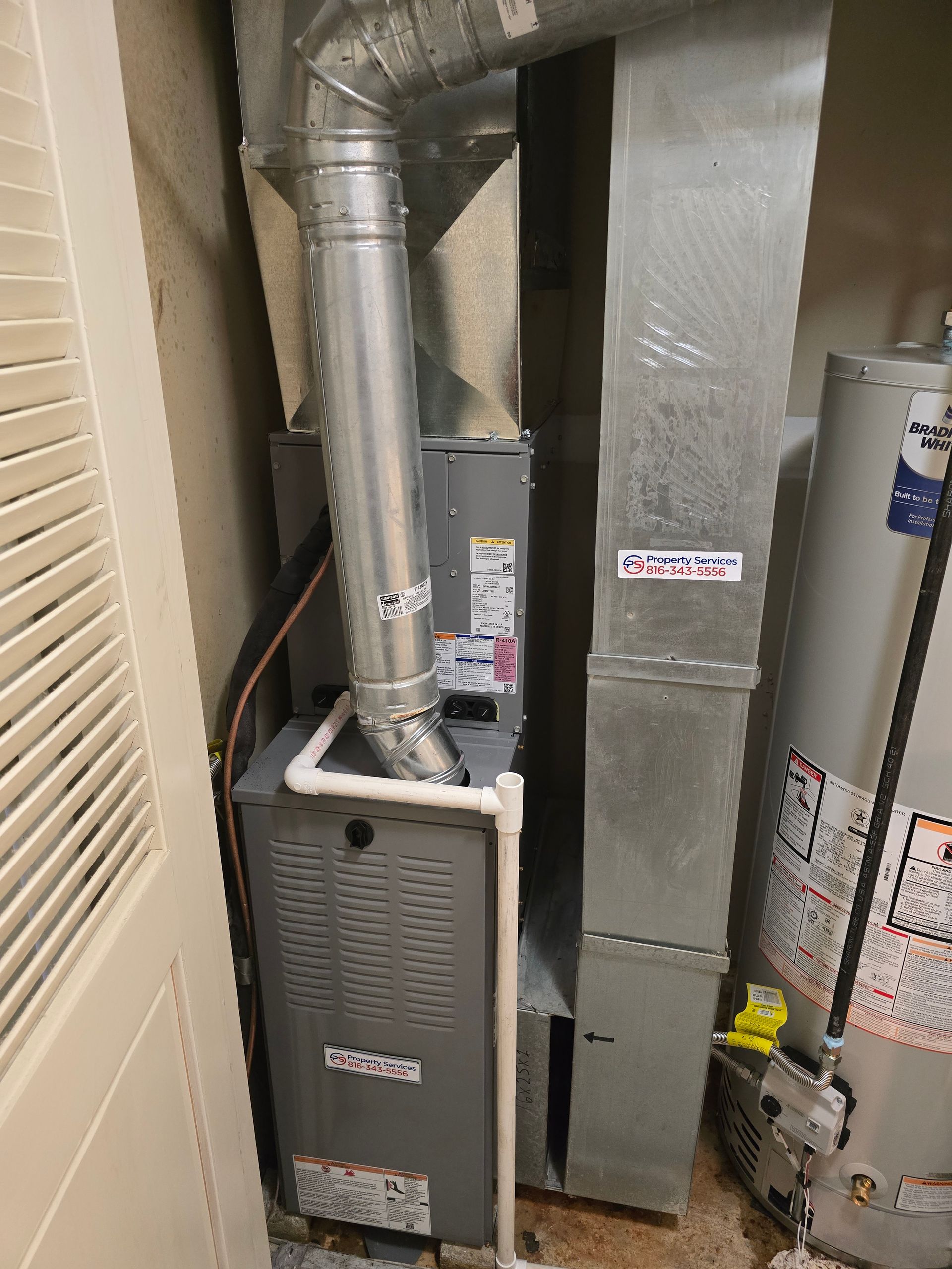 A gray furnace with white PVC piping connected to silver ductwork next to a water heater in a utility closet.