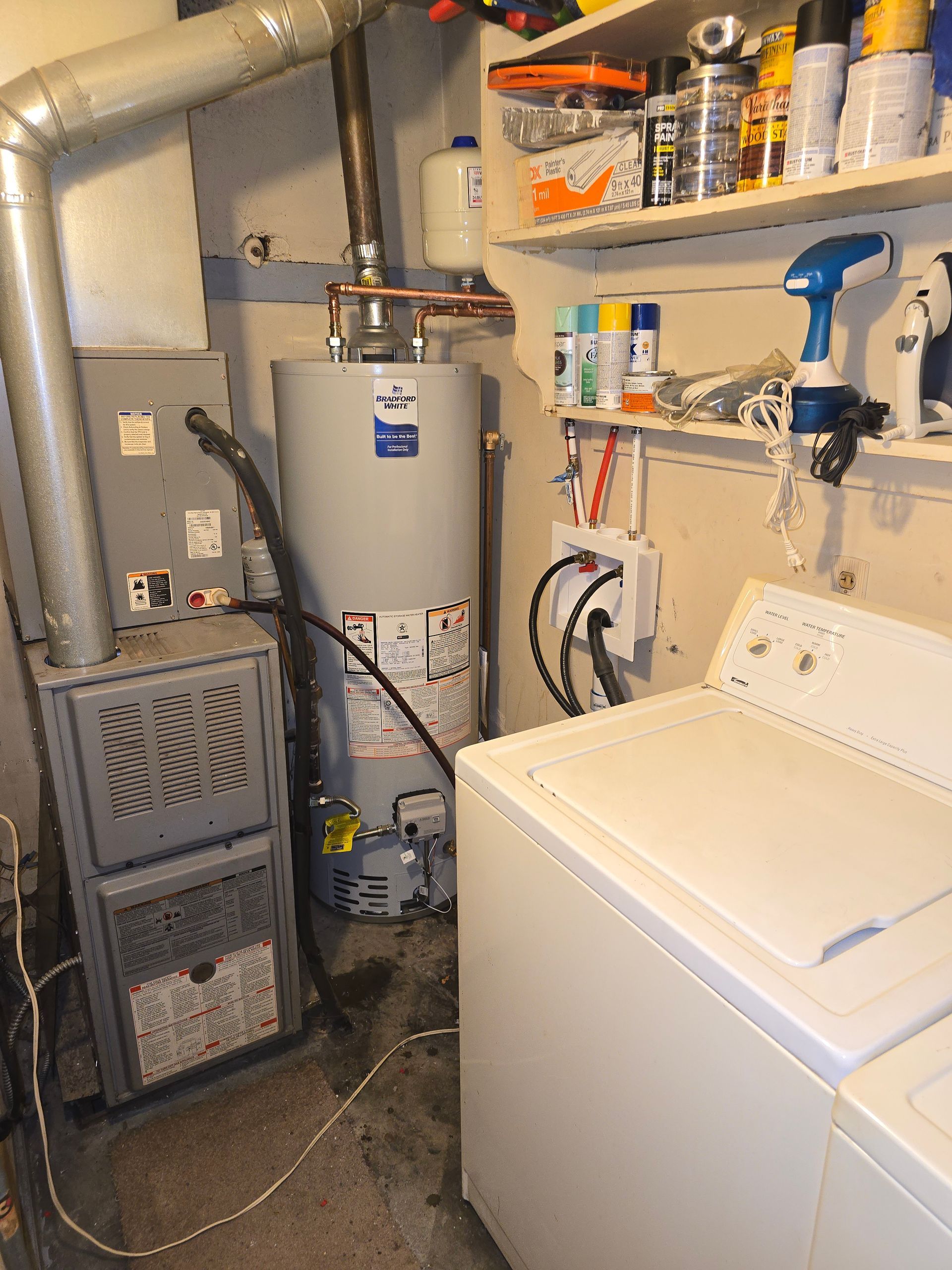 A furnace and a water heater stand in a utility room next to a white washing machine and wall-mounted storage shelves.