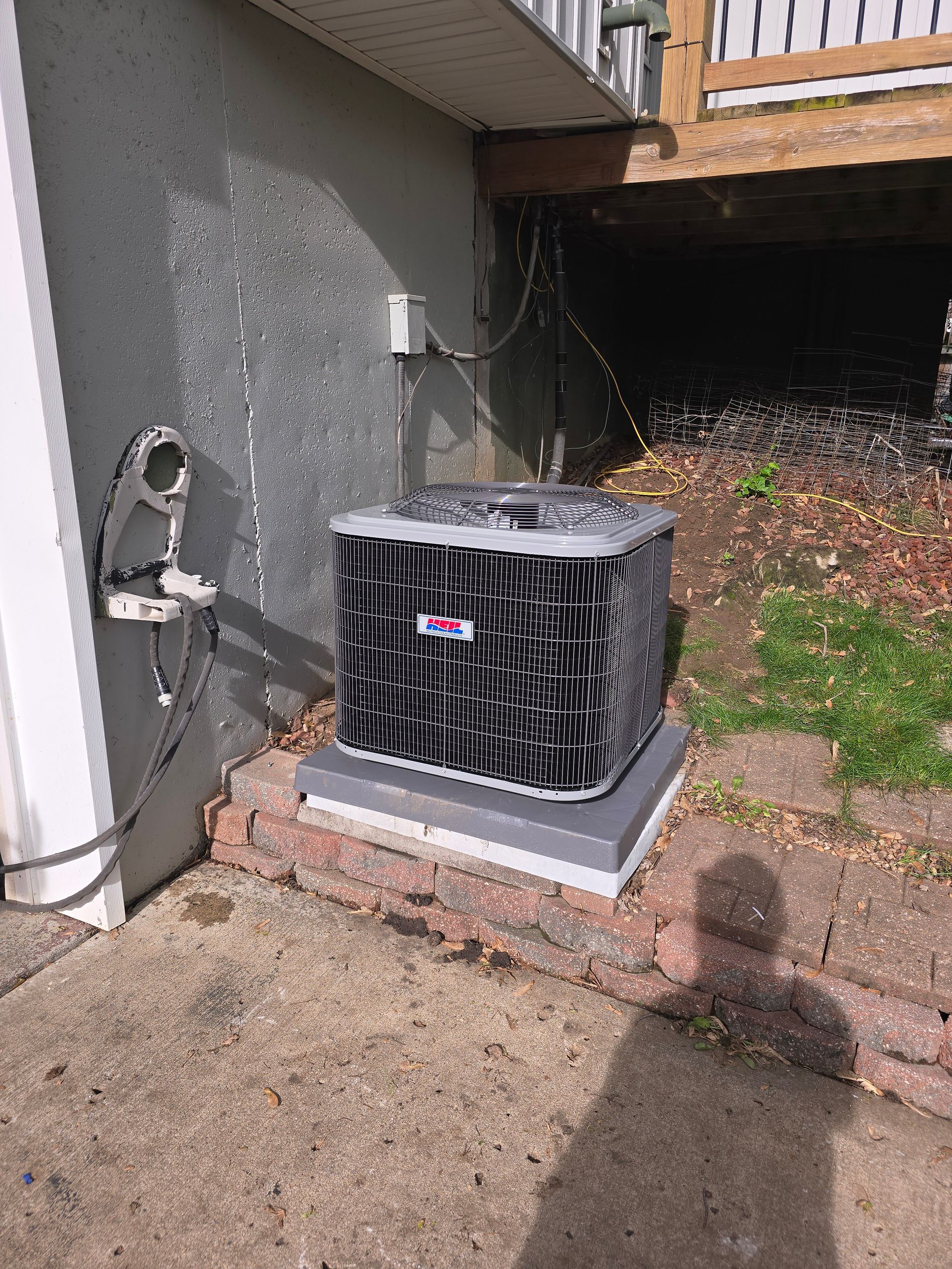 An outdoor air conditioning unit sits on a concrete pad near a house wall under a wooden deck.
