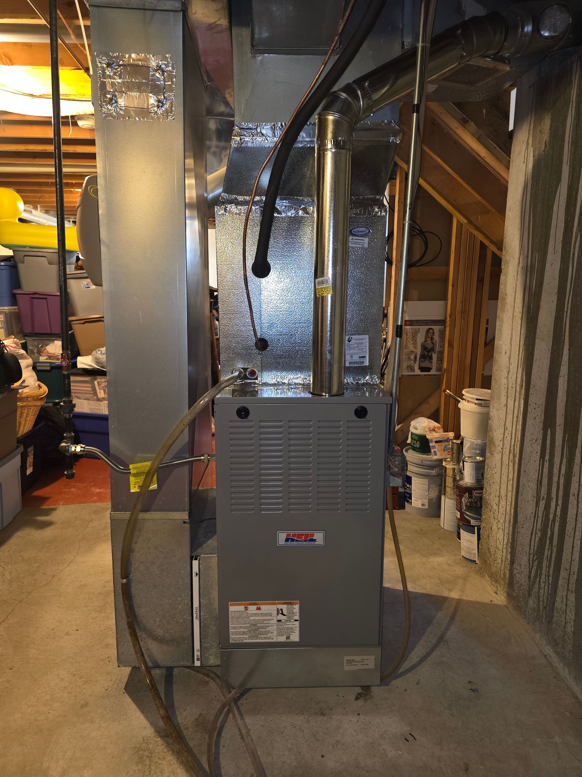 A gray natural gas furnace installed in a basement, connected to vertical metal ductwork and a venting pipe.