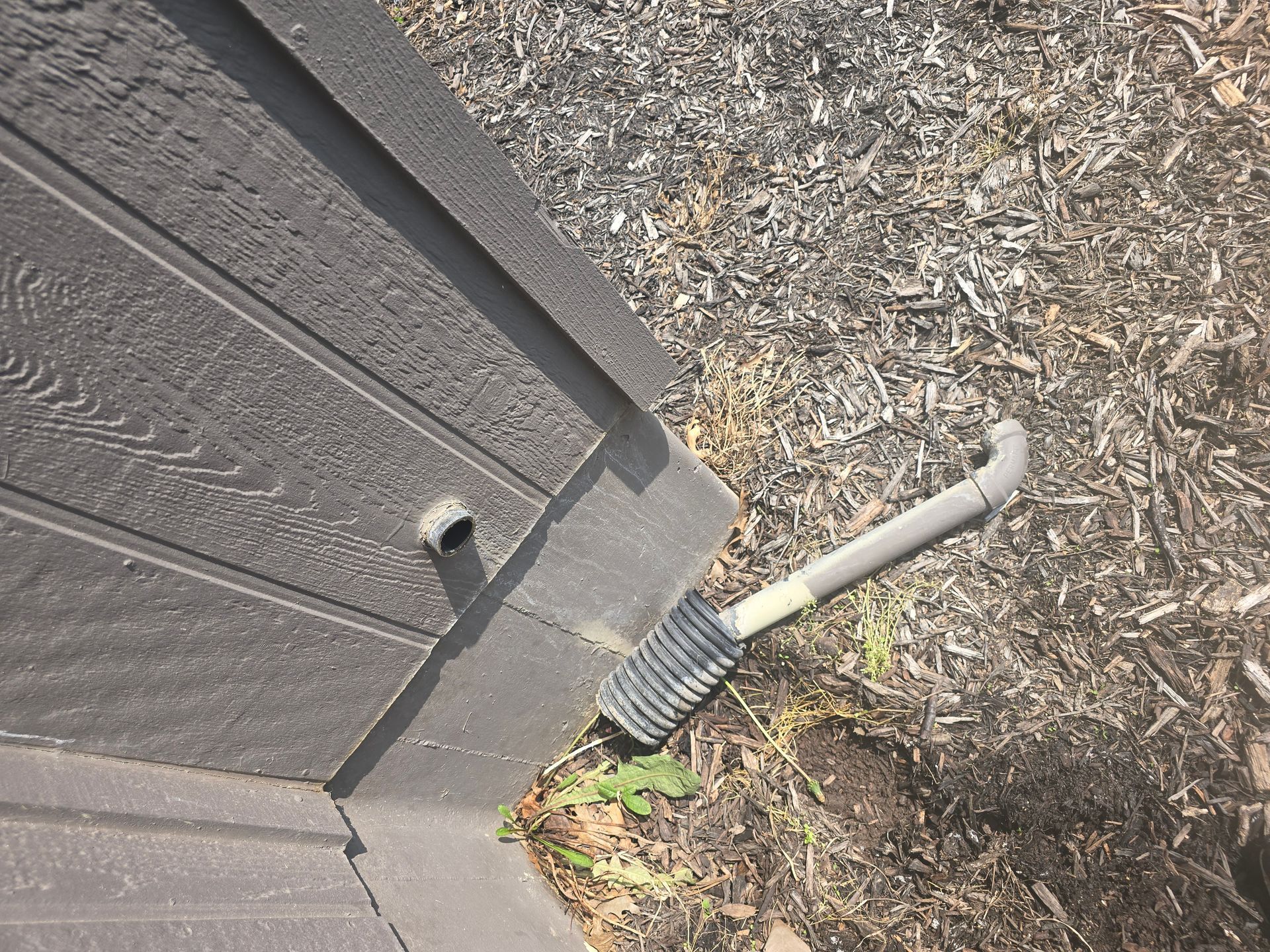 A grey drainage pipe protruding from a shed wall sits next to a corrugated flexible pipe resting on dark mulch.