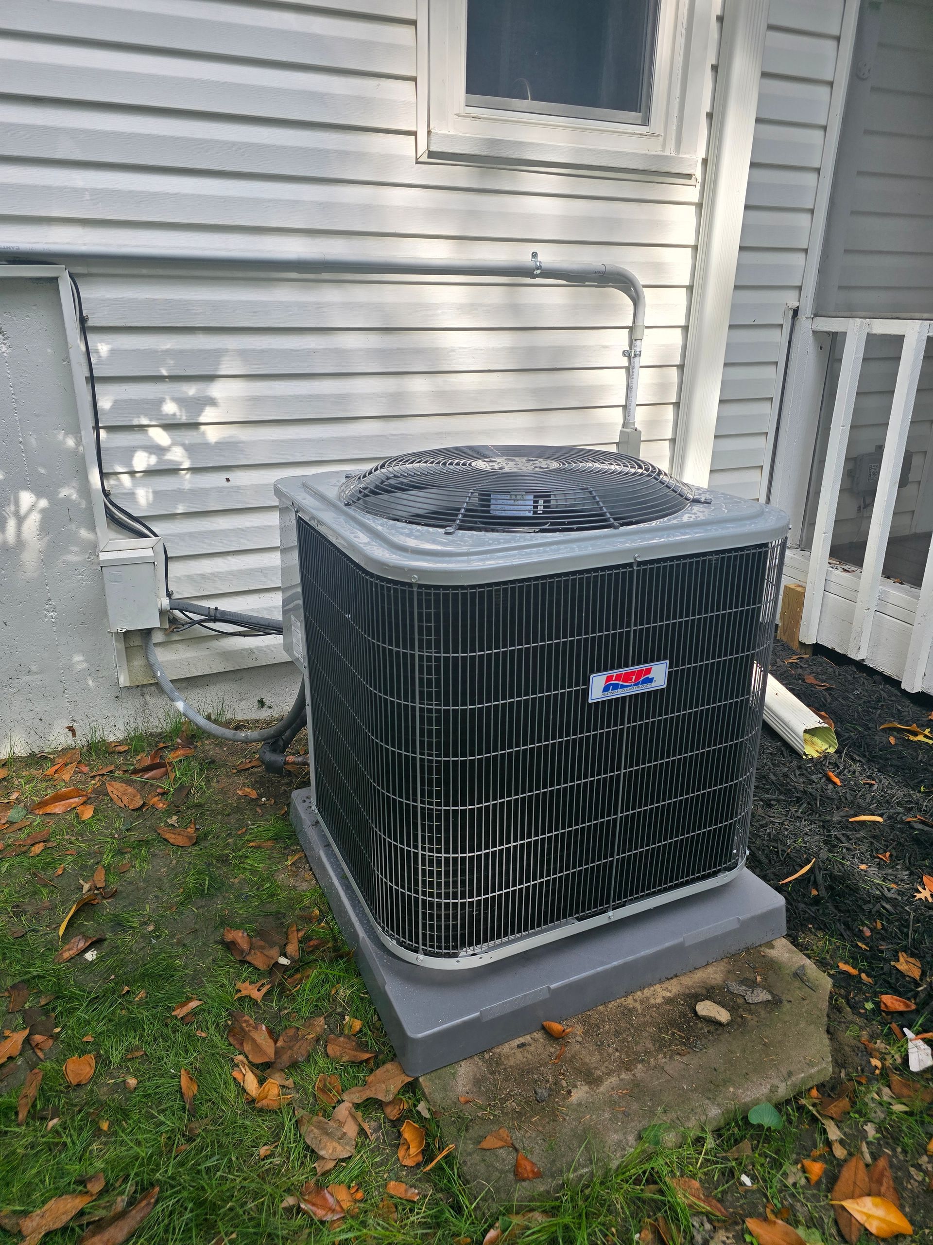 A gray residential air conditioning unit sits on a concrete pad outside a house with white siding.