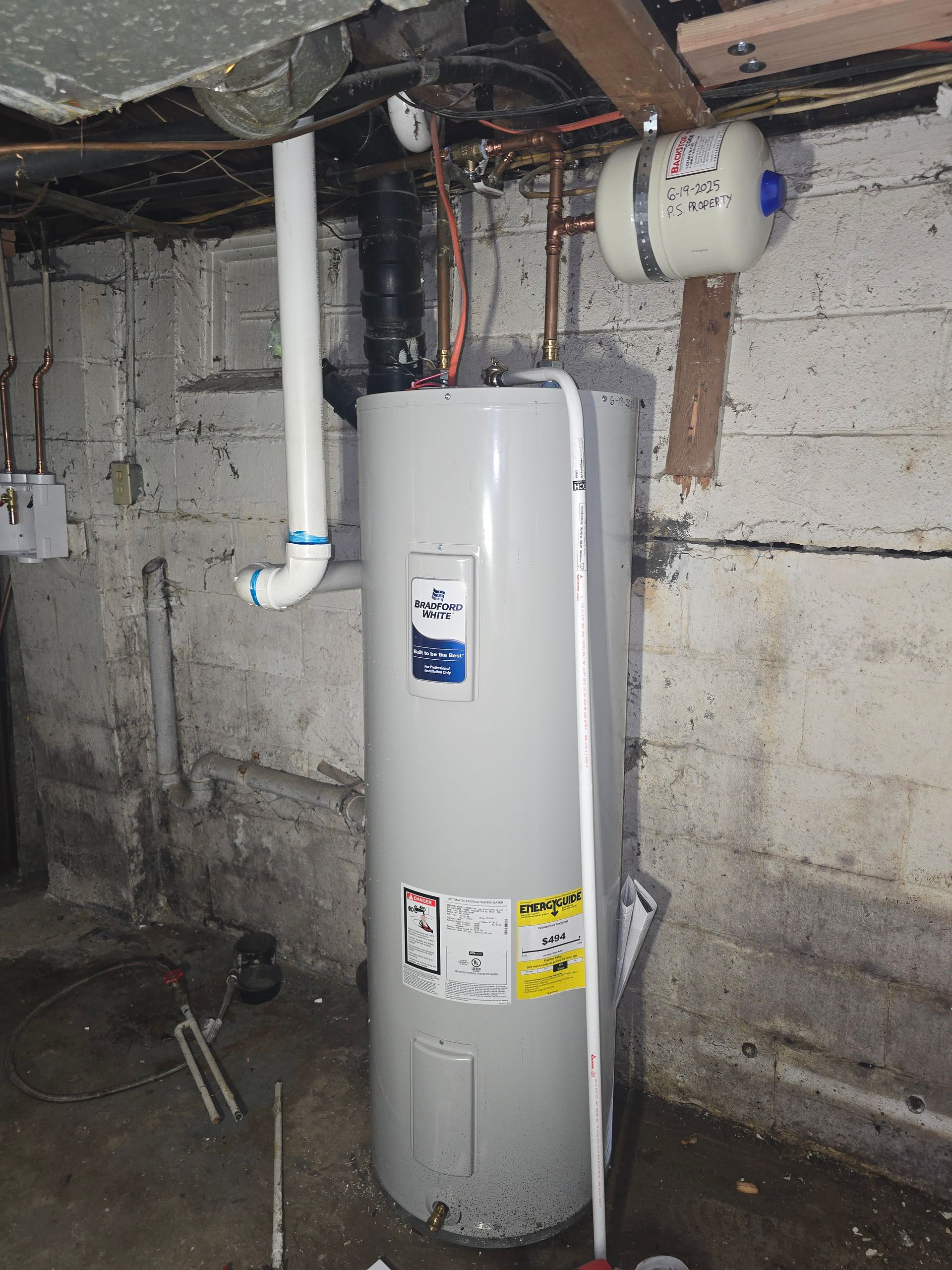 A grey water heater in a basement with exposed copper pipes and a small white expansion tank mounted above.