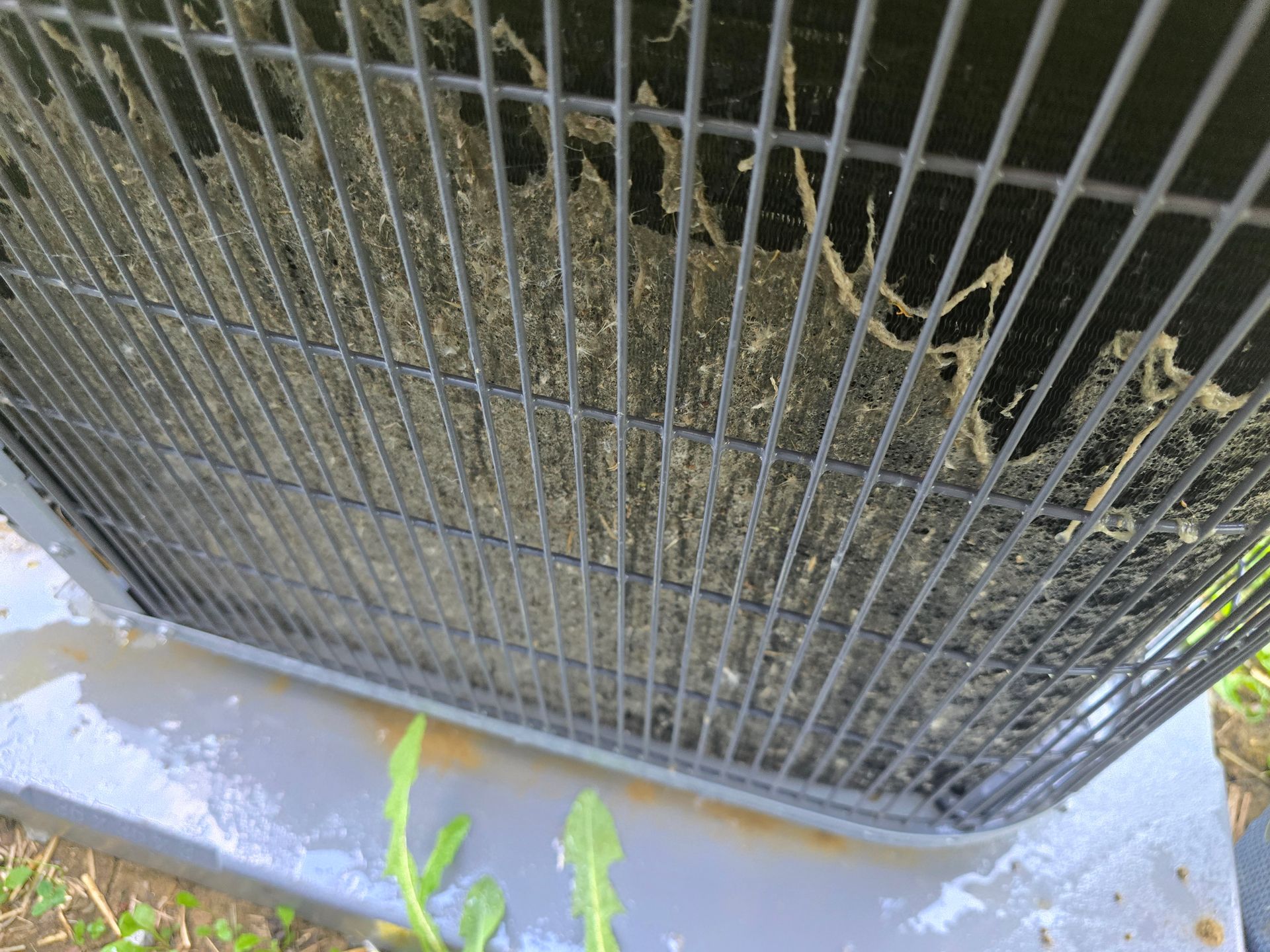 The metal fins of an outdoor air conditioning unit are heavily coated with thick, matted gray debris and dust.