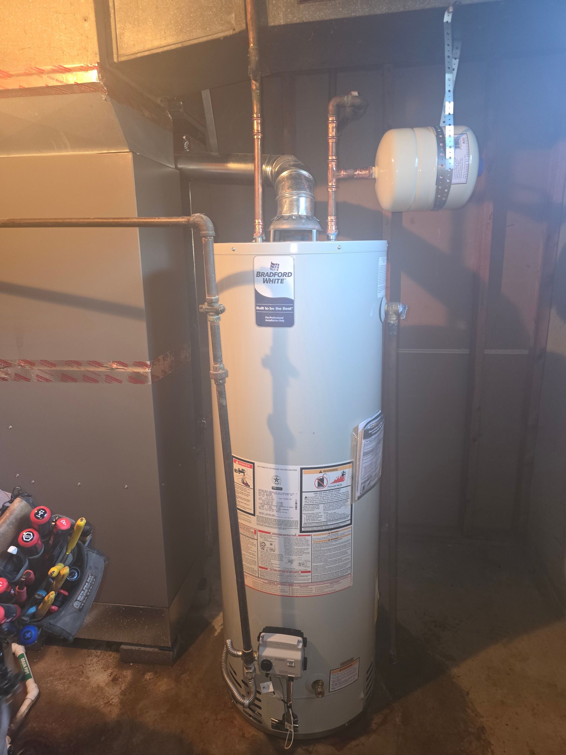 A light gray residential water heater stands in a basement with connected copper plumbing and a small expansion tank.