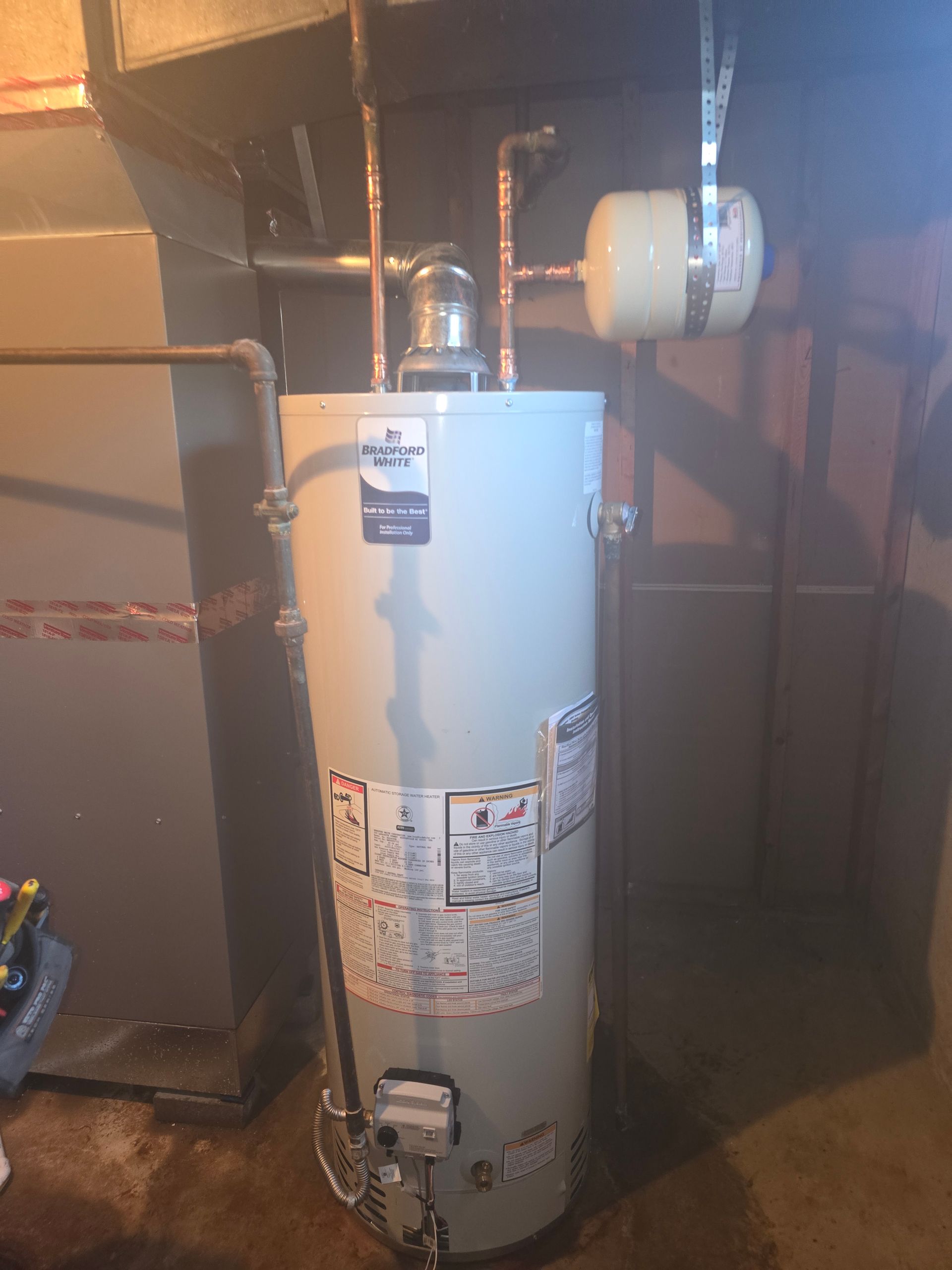 A beige residential water heater installed in a basement, featuring copper piping and an attached expansion tank.