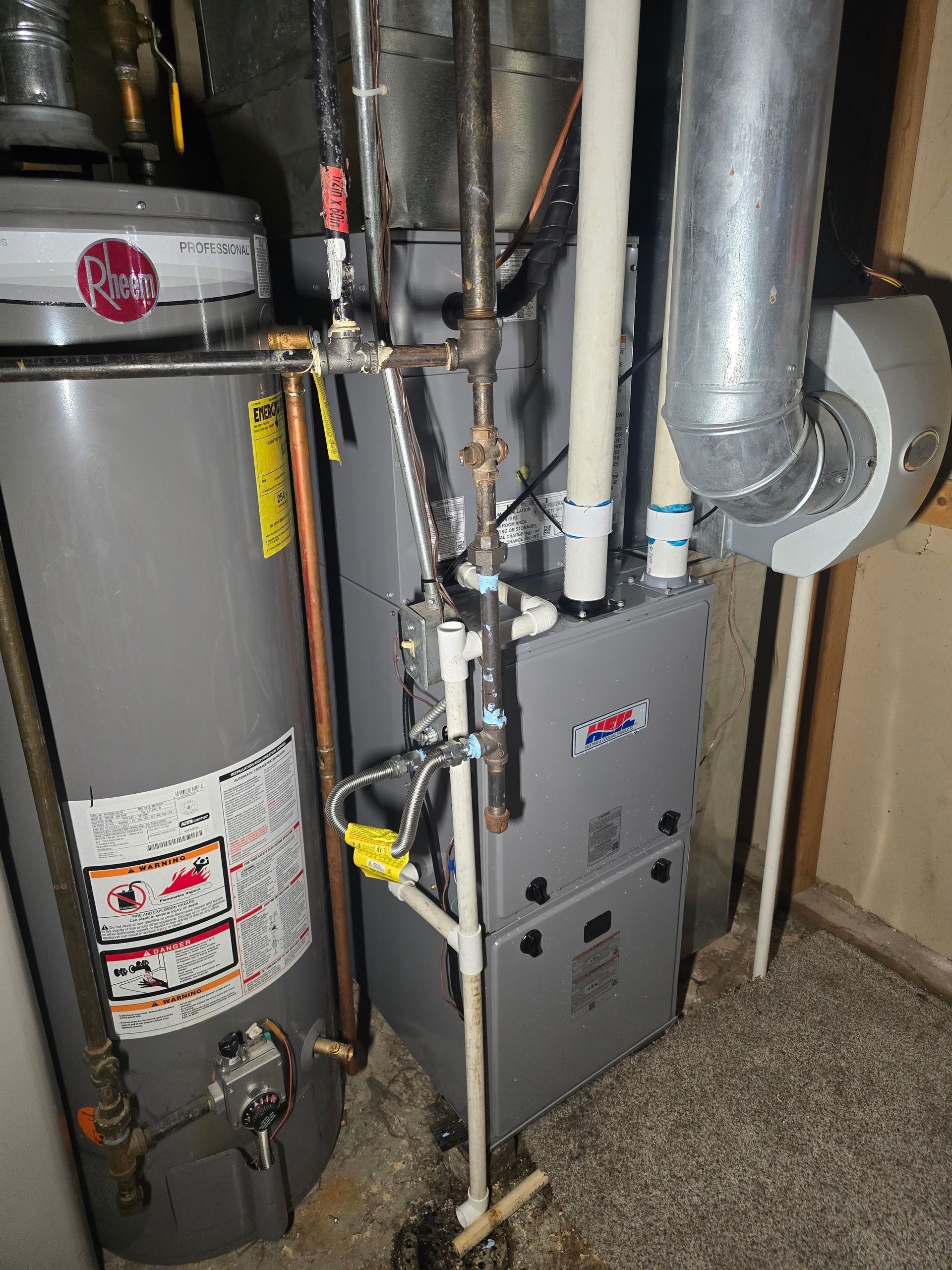 A Rheem water heater beside a grey gas furnace with white PVC venting pipes in a basement mechanical room.