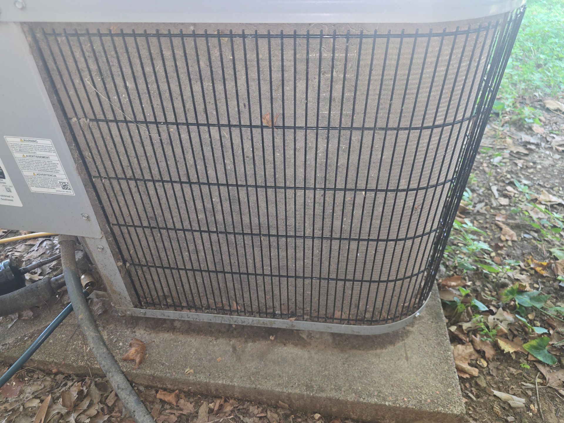 An outdoor air conditioning unit with a black protective grille, sitting on a concrete slab near leaves and dirt.