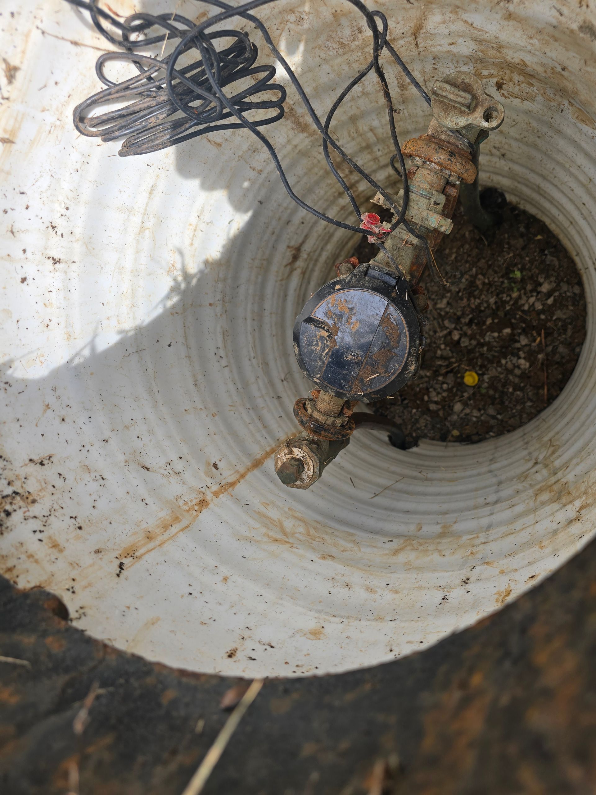 A water meter inside a white, cylindrical underground utility box, with loose black wires resting on top of the meter.