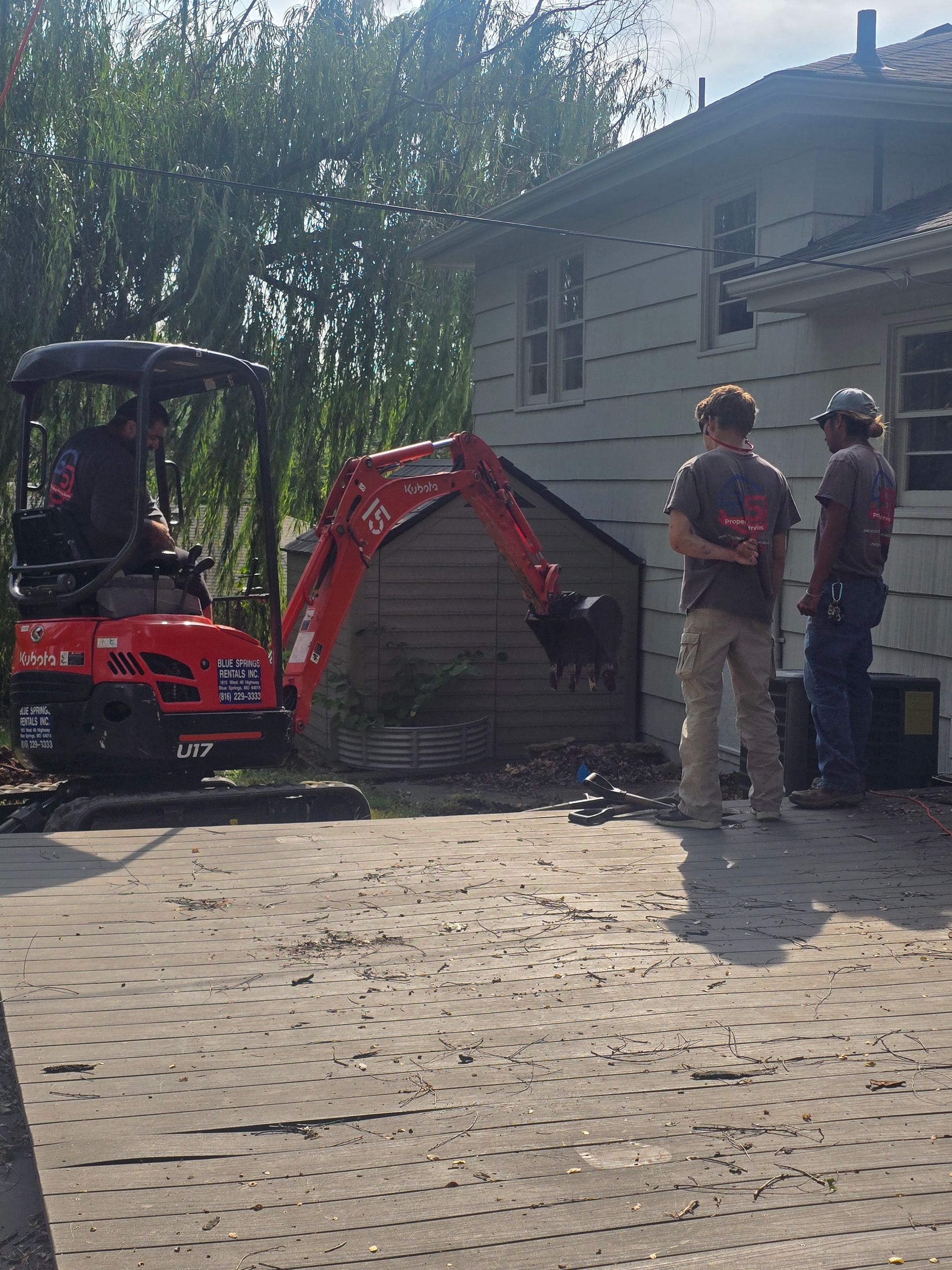 Two people stand outside a house watching a bright red mini excavator operating near the building's exterior wall.