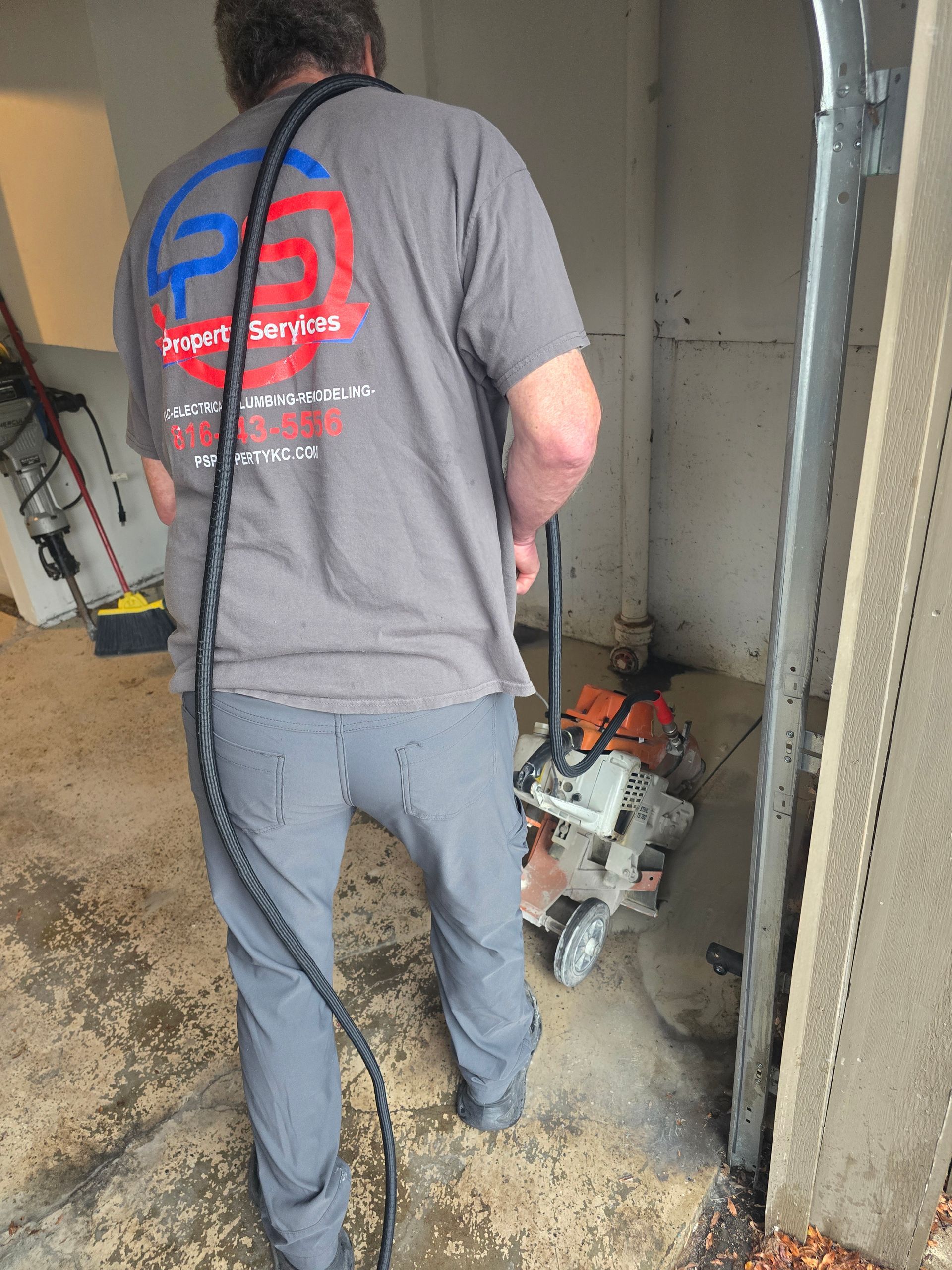 A worker in a gray uniform uses a concrete saw to cut a floor in an indoor space.