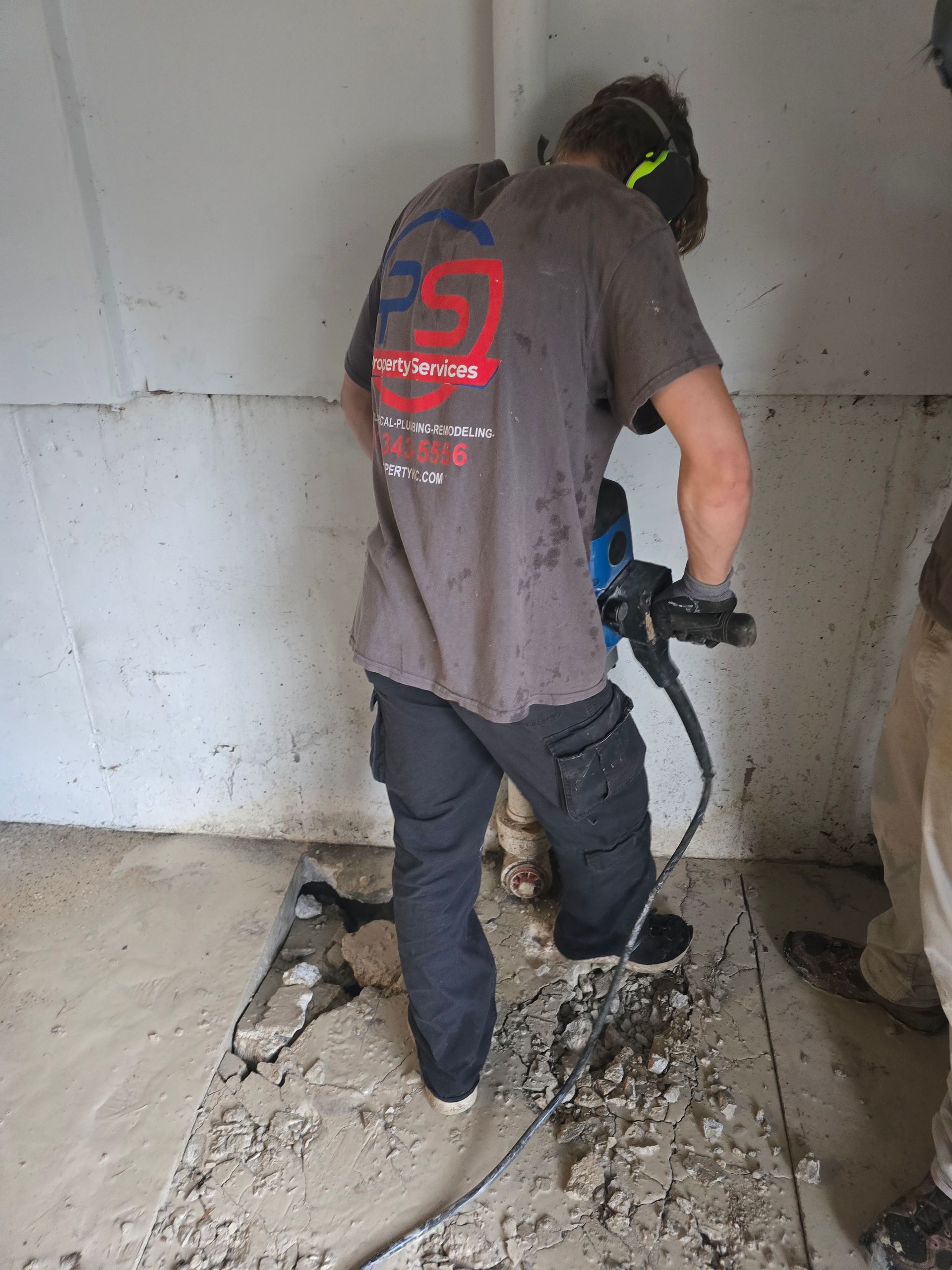 A worker in a gray shirt uses a jackhammer to break up a concrete floor indoors.