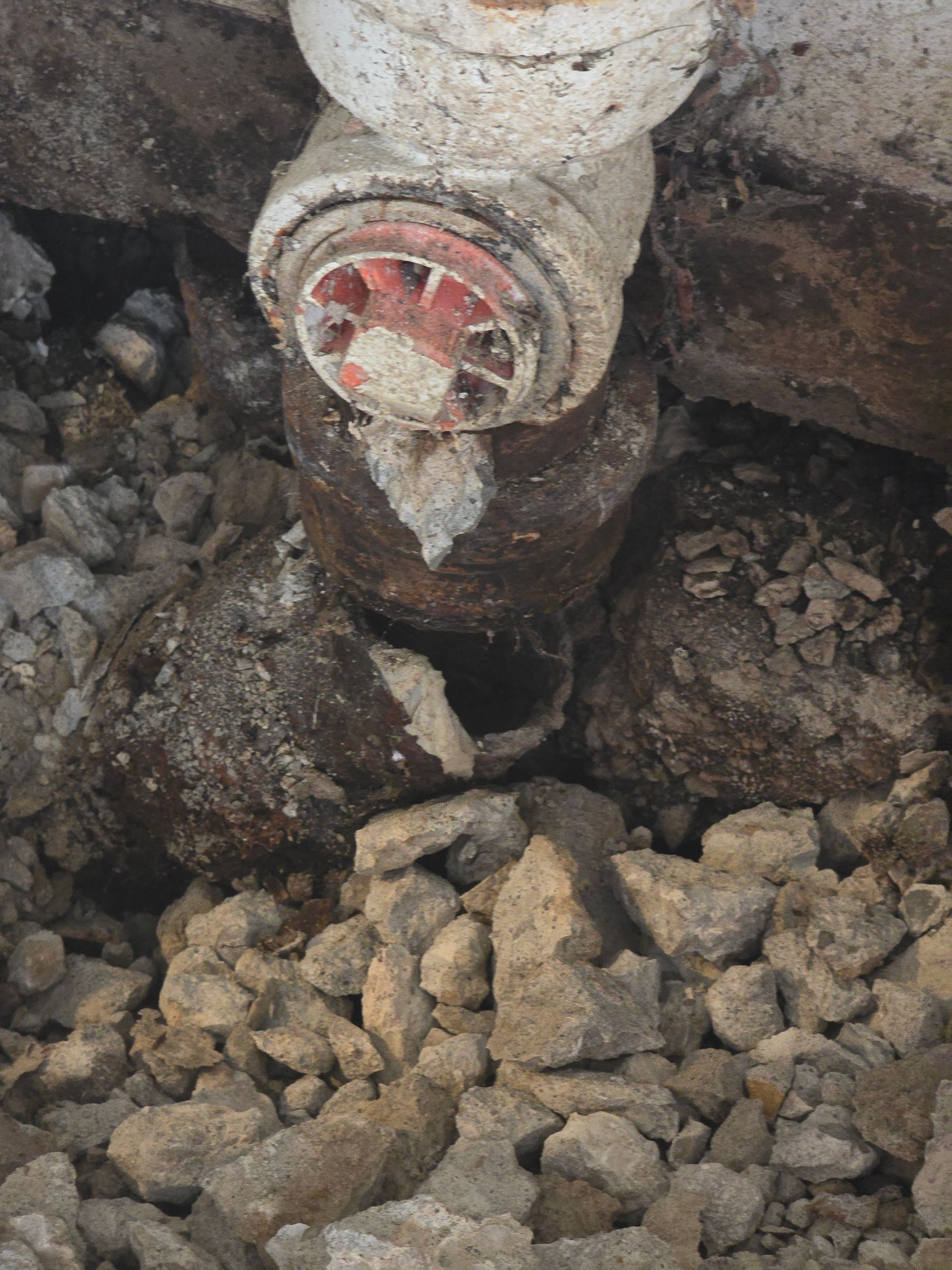 A damaged, rusted pipe connection showing a clear hole in the side, situated in a hole filled with loose gravel and dirt.