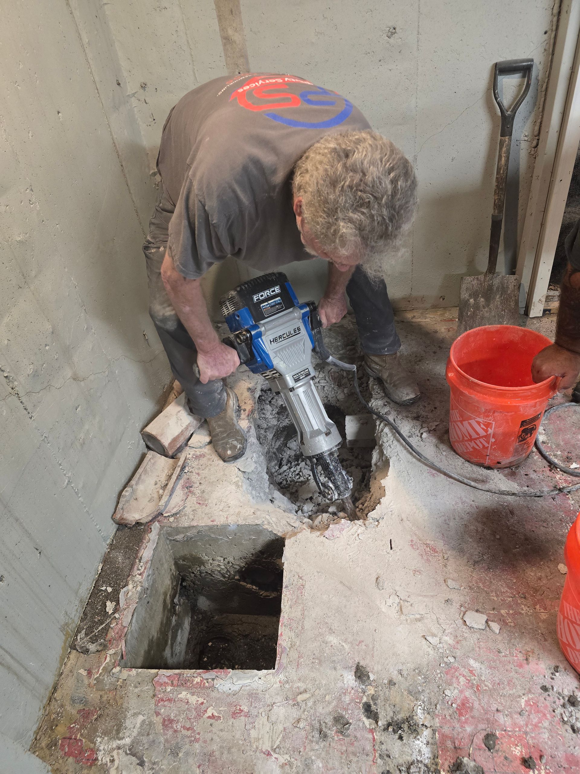 A person uses a jackhammer to break through a concrete floor in an indoor space near a bucket and a shovel.