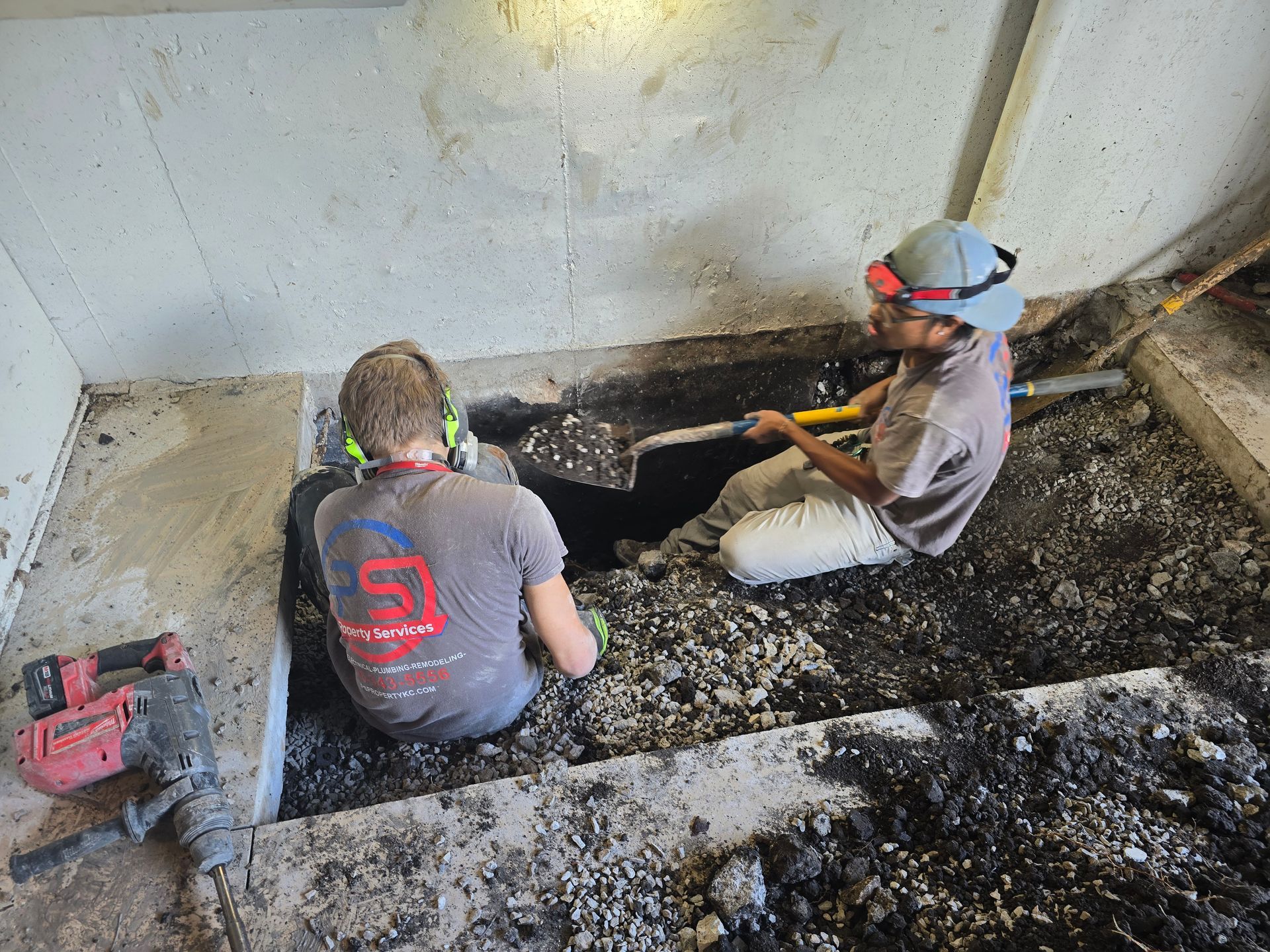 Two workers use shovels to excavate soil from a trench cut into a concrete floor, with a jackhammer nearby.