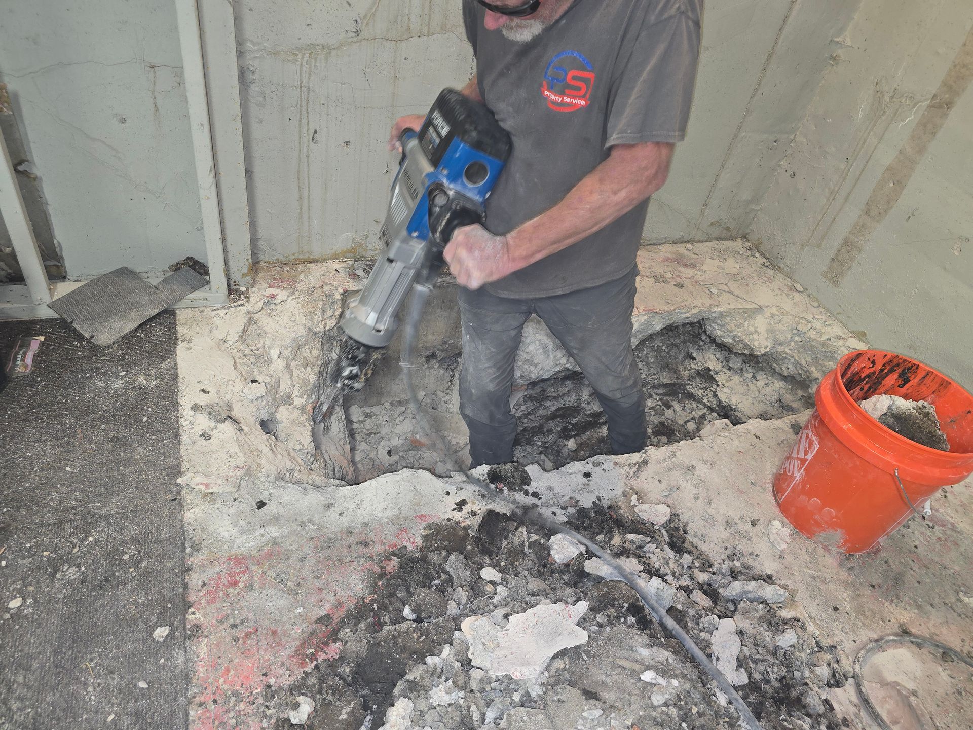 A person uses a jackhammer to break up a concrete floor in an indoor construction site near an orange bucket.