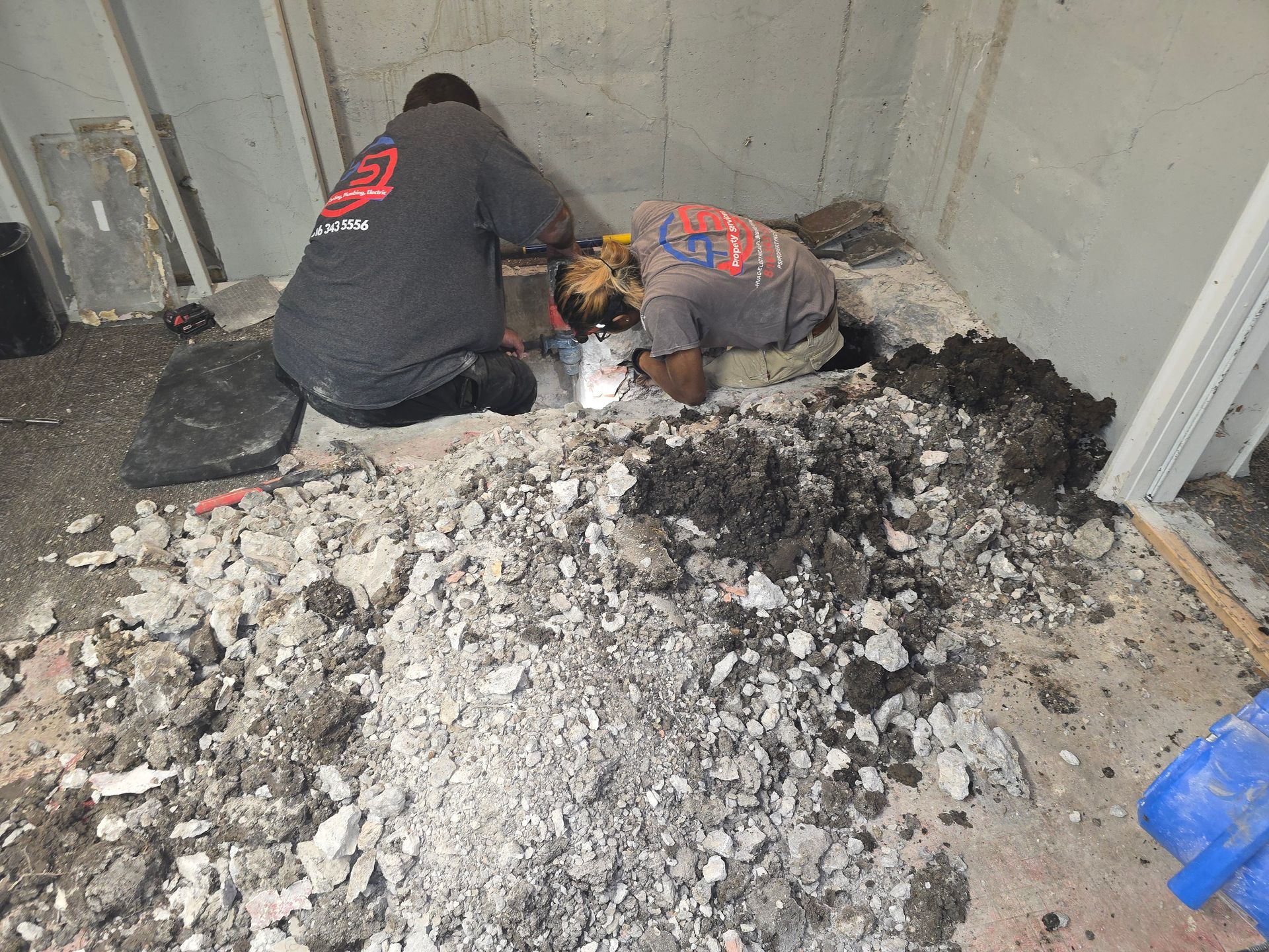 Two workers in dark shirts kneel on a basement floor, breaking up concrete to access a hole in the ground.