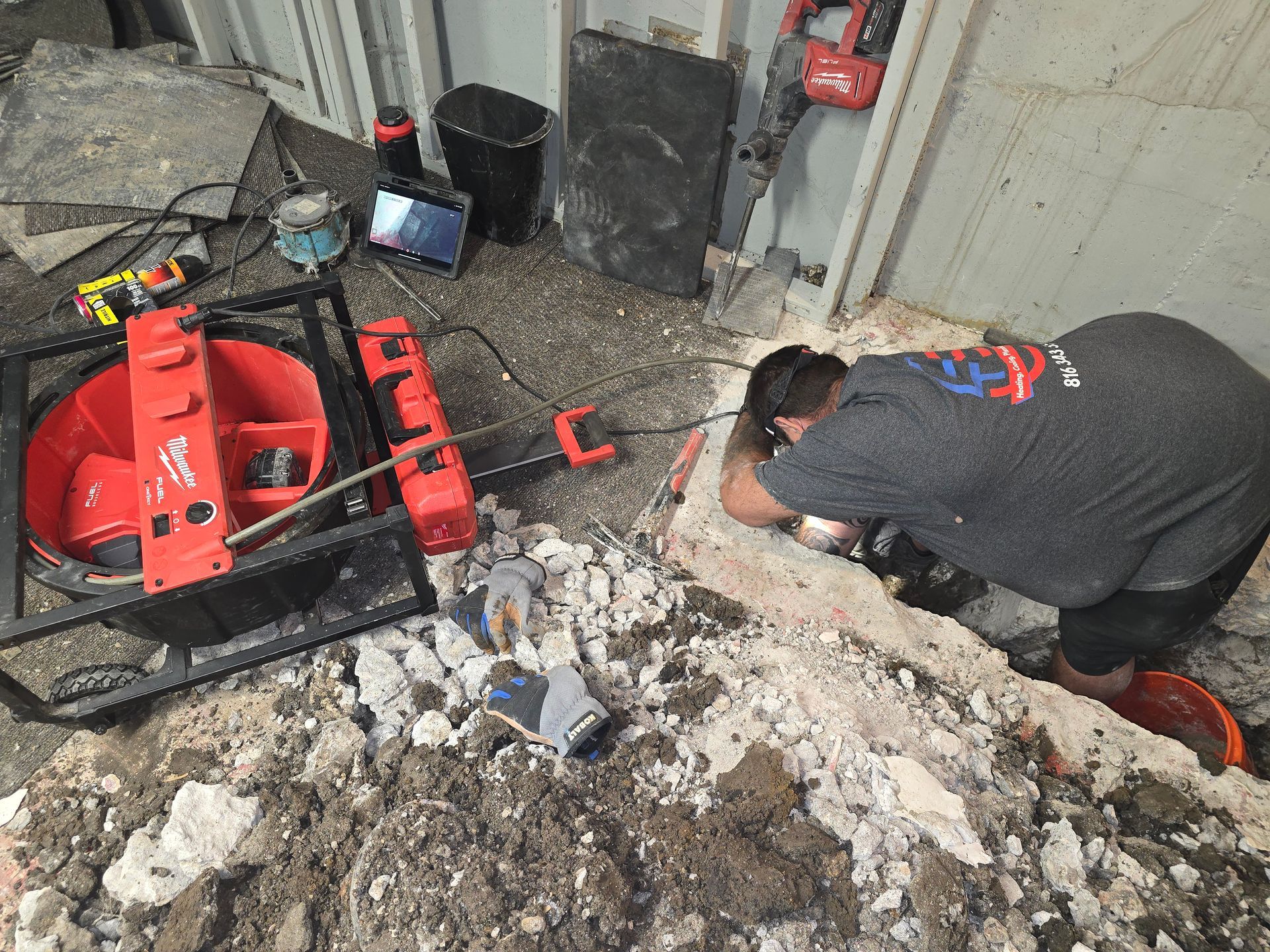 A worker in a gray t-shirt inspects underground plumbing pipes in a dirt-filled trench near a red Milwaukee tool unit.
