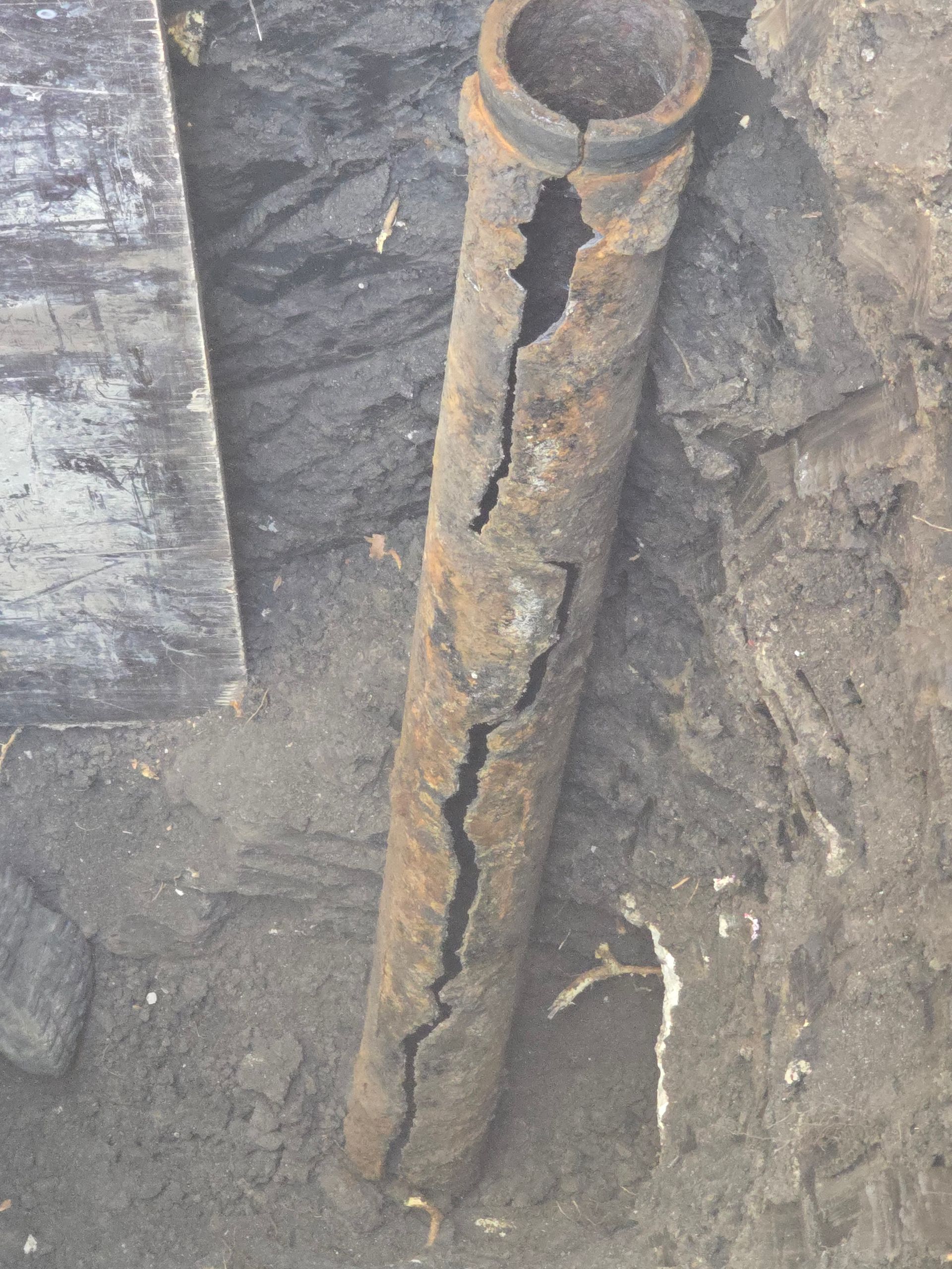 A deeply corroded, rusted metal pipe with a large vertical crack, partially buried in dirt.