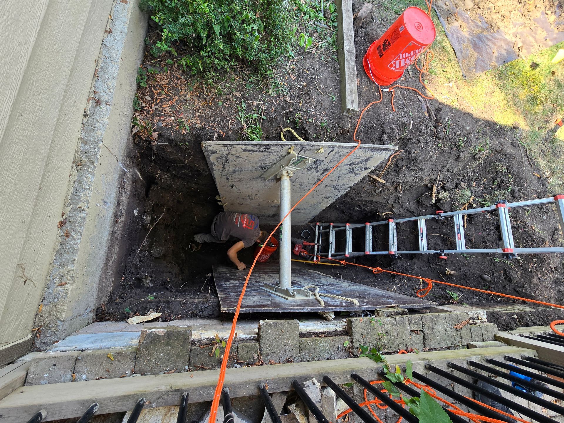 A worker in an excavated trench beside a building, supported by a metal post and planks, with a ladder for access.