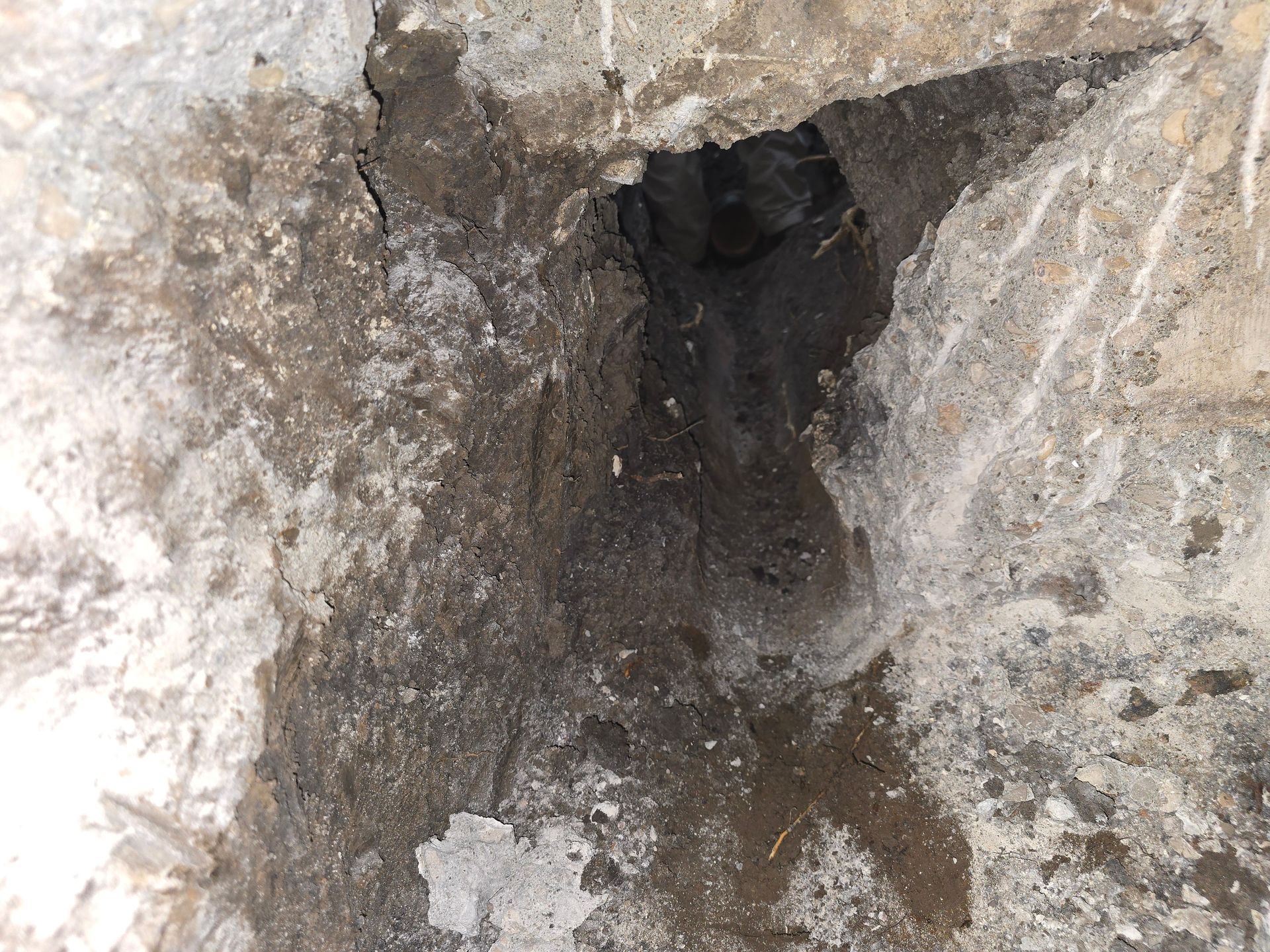 A jagged, dark hole broken into a rough, light-colored concrete or stone surface, revealing dirt underneath.