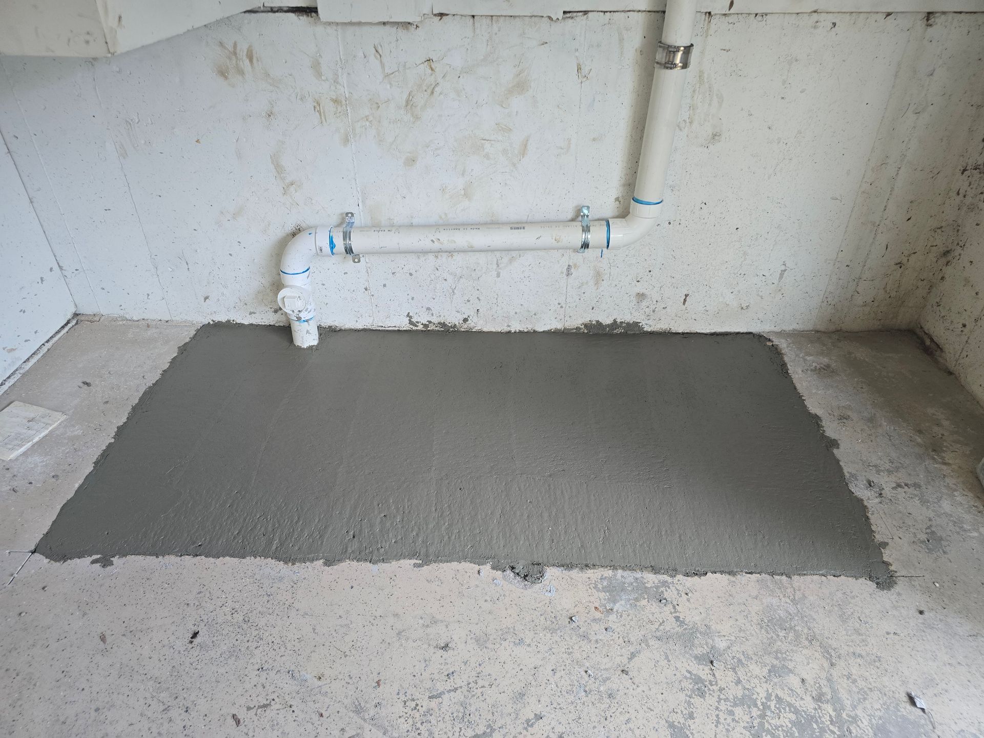 Freshly poured concrete patch on a basement floor beneath white PVC plumbing pipes against a concrete wall.