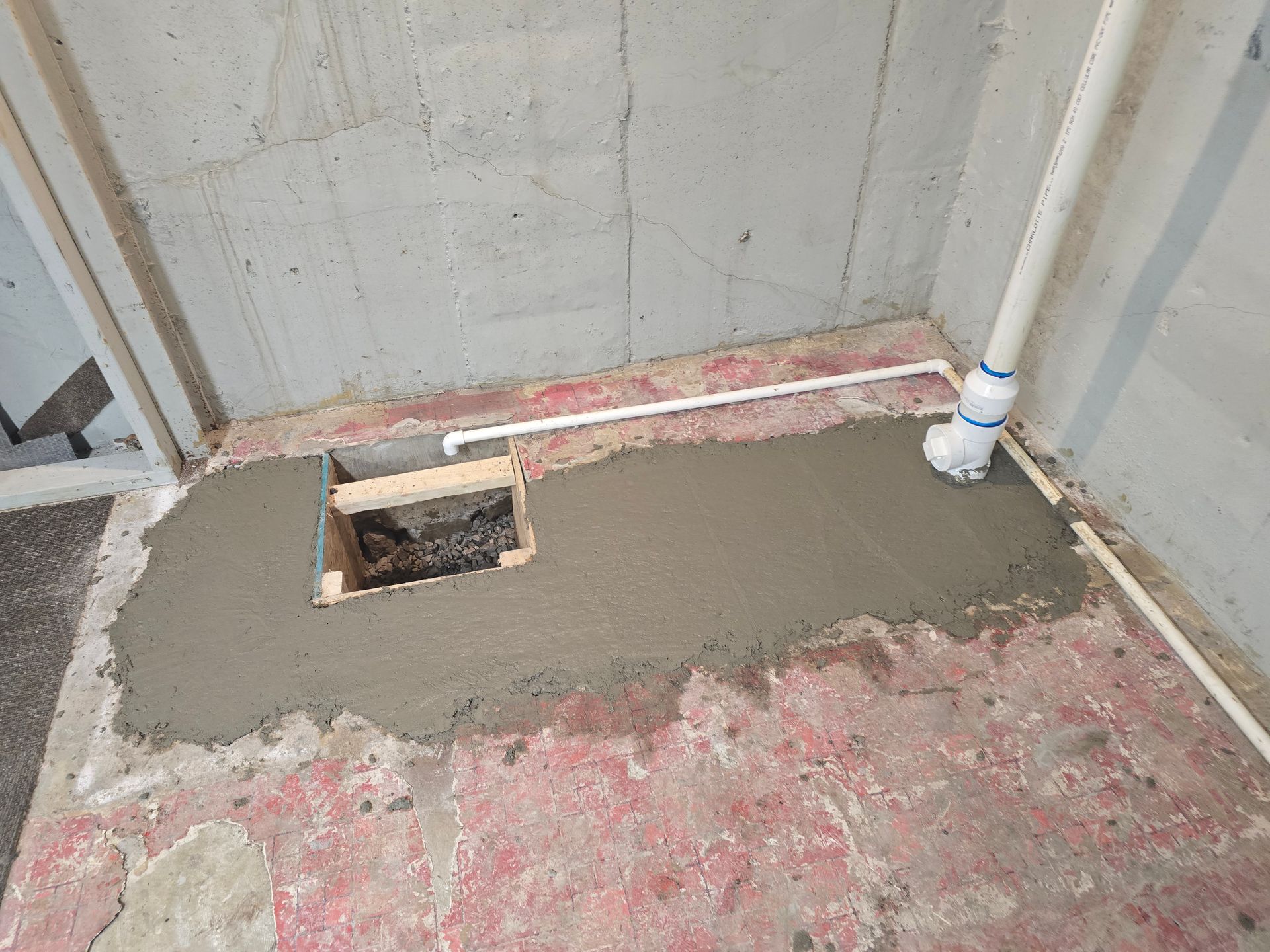 A concrete patch fills part of a basement floor, surrounding a wooden framed opening and a white PVC pipe.