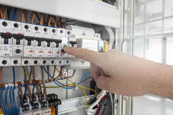 Hand pointing at electrical breakers inside an open control panel with wiring.