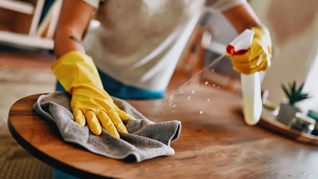 A person wearing yellow gloves is cleaning a wooden table with a cloth and spray bottle.