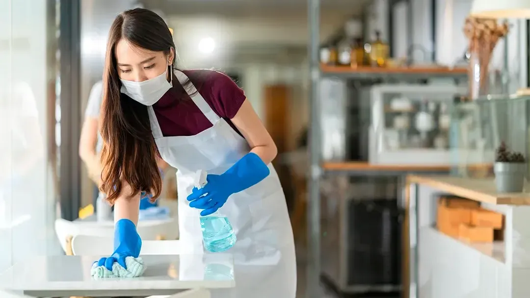 A woman wearing a mask and gloves is cleaning a table in a kitchen.