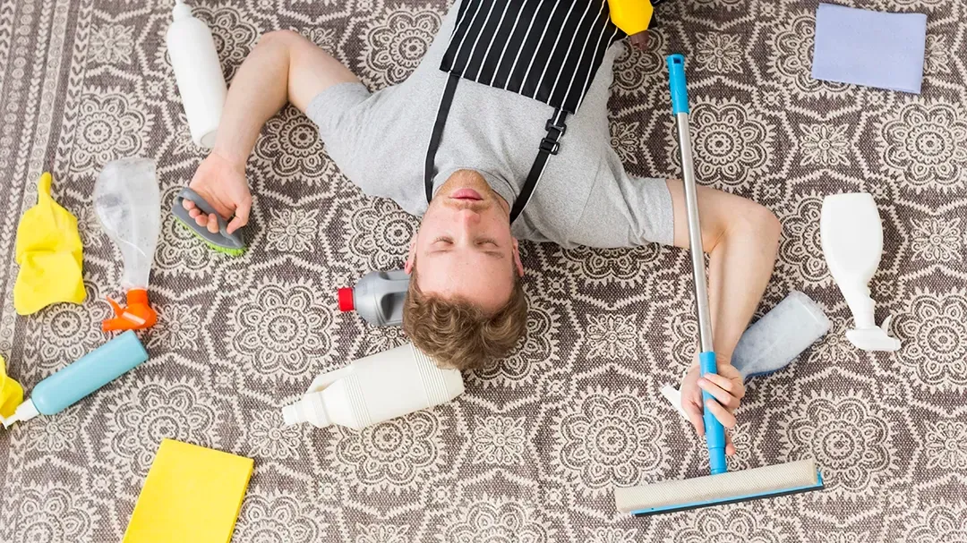 A man is lying on the carpet floor surrounded by cleaning supplies.