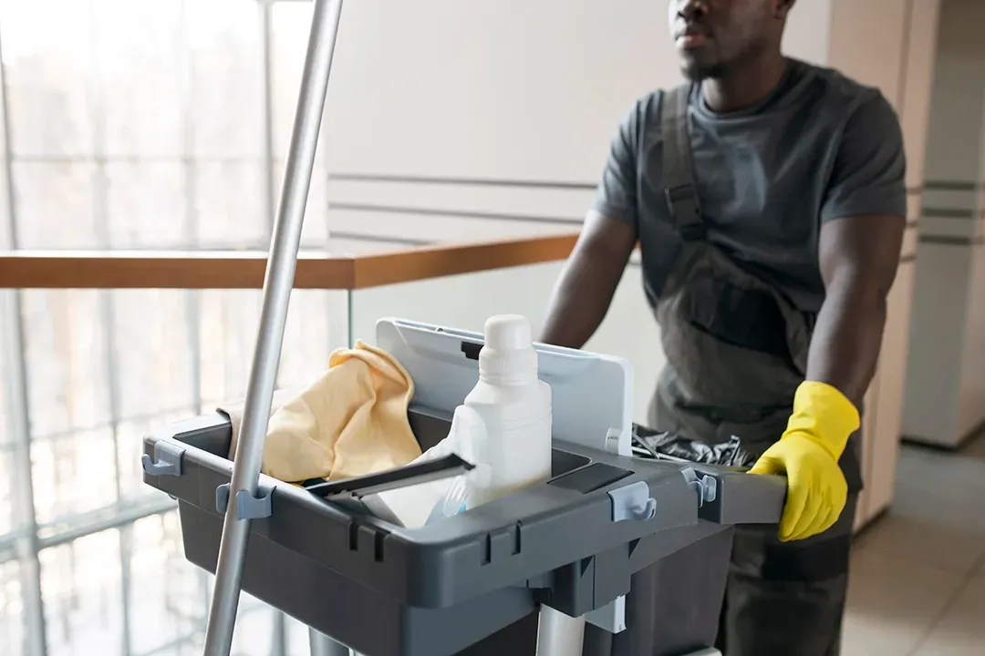 A man wearing yellow gloves is pushing a cart with cleaning supplies.