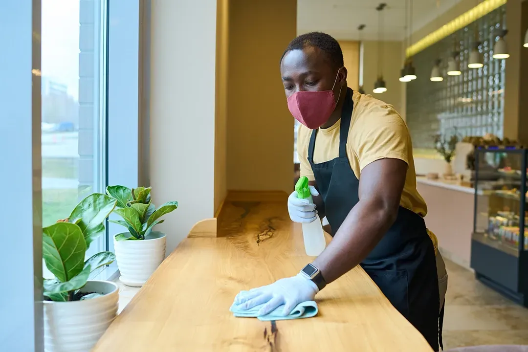 A man wearing a mask is cleaning a counter in a restaurant.