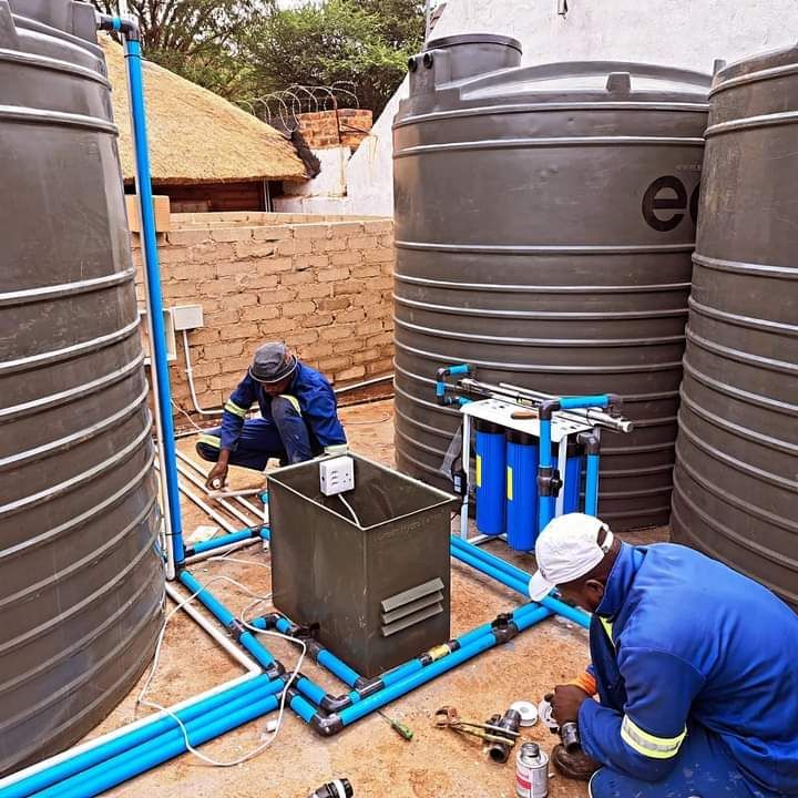 Two men are working on a water system in front of large tanks with the letter e on them