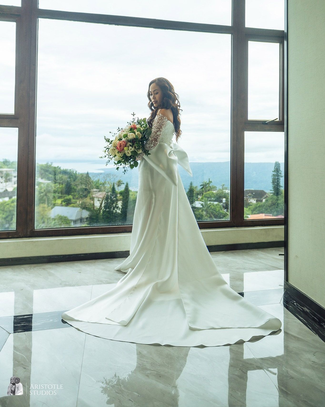 A woman in a wedding dress is standing in front of a window holding a bouquet of flowers.