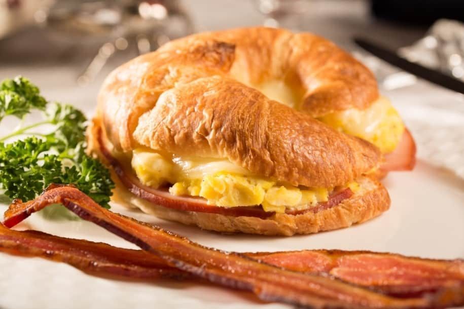 A breakfast sandwich on a croissant with bacon and eggs on a plate.