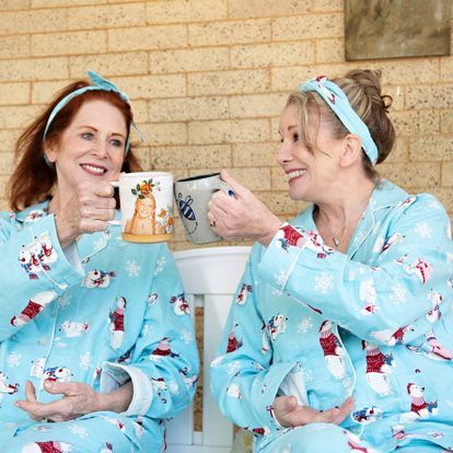 Two women in pajamas are sitting on a bench drinking coffee