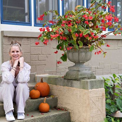 A woman is sitting on the steps of a house next to pumpkins and flowers.