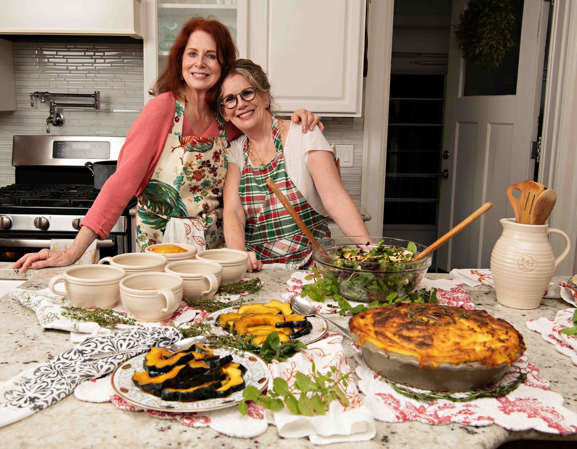 Two women are posing for a picture in a kitchen with food on the table.