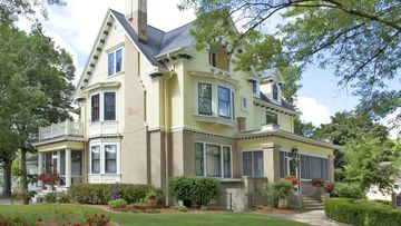 A large yellow house with a black roof is surrounded by trees and bushes.