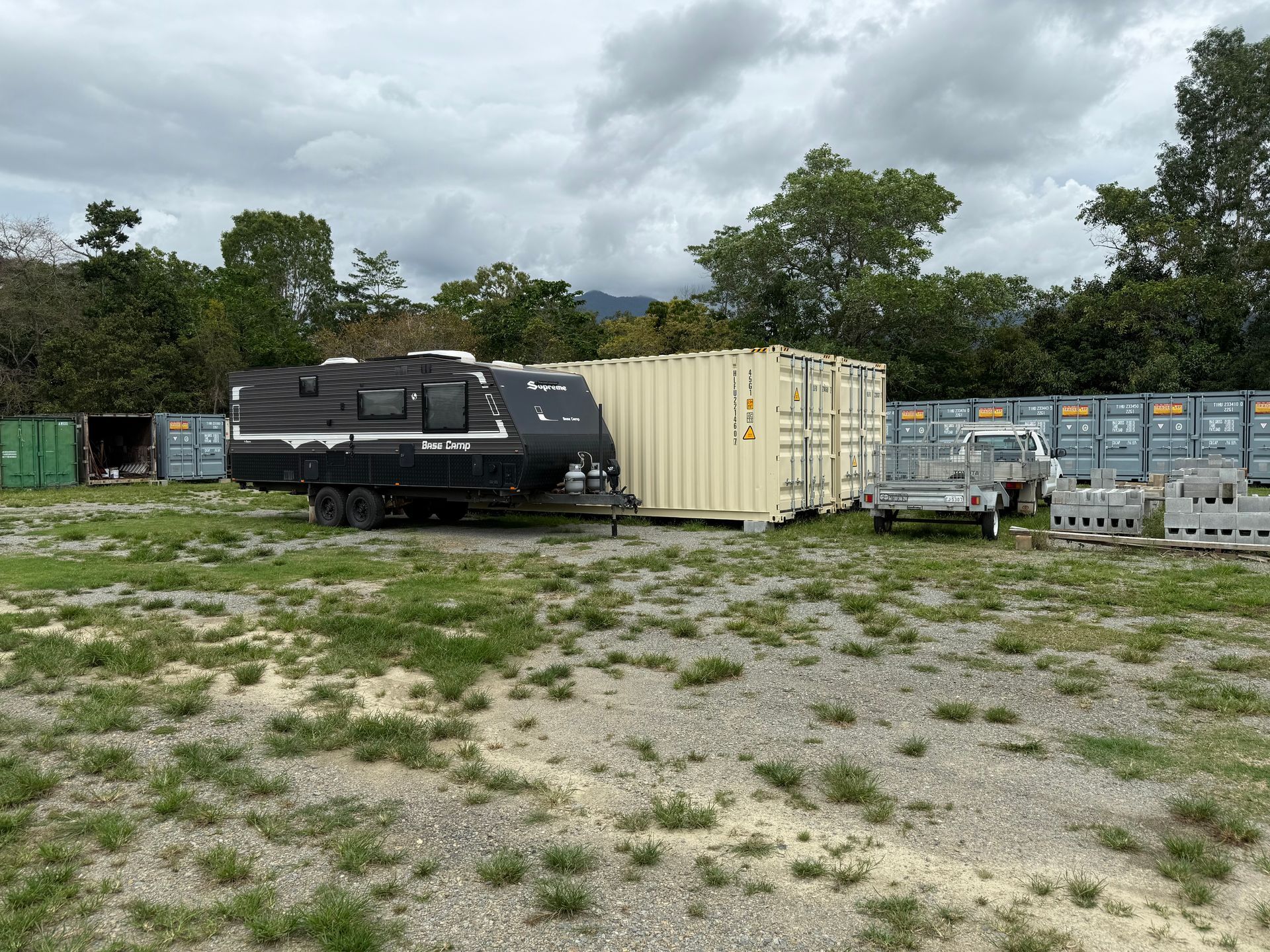 A dark travel trailer parked next to a beige shipping container on a grassy lot with other containers and trees — Cairns Budget Storage In Mount Sheridan, QLD