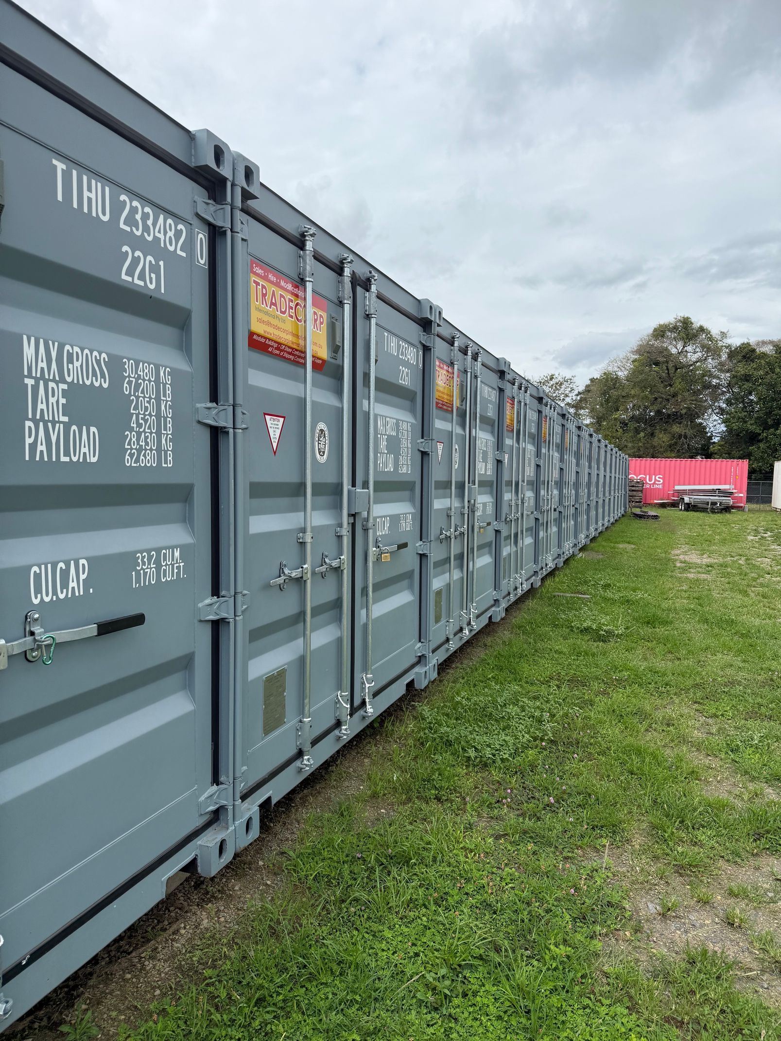Row of gray shipping containers in a grassy field, likely for storage, with a cloudy sky overhead.— Cairns Budget Storage In Mount Sheridan, QLD